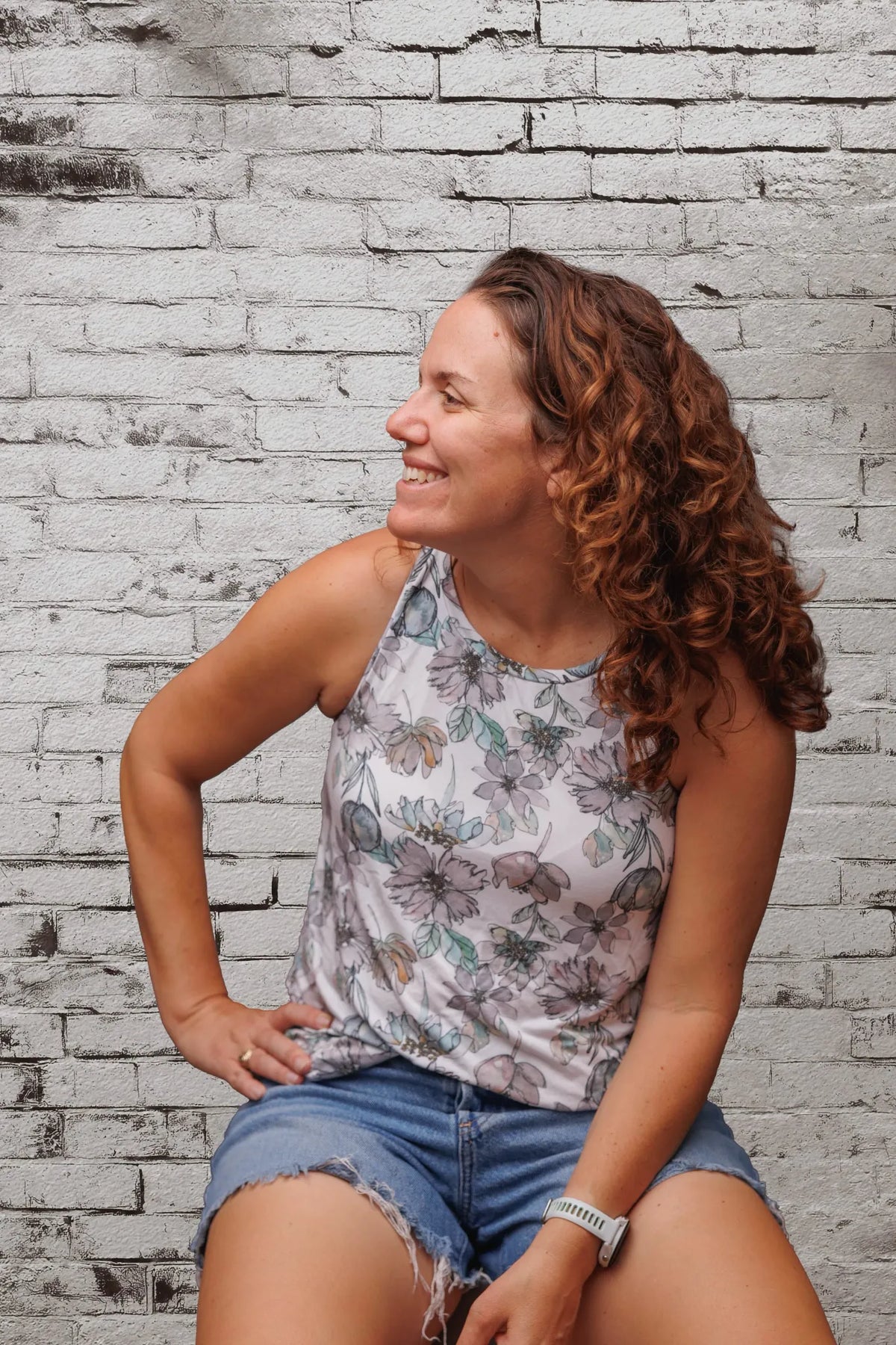 A woman in a floral top and denim shorts sits against a white brick wall.