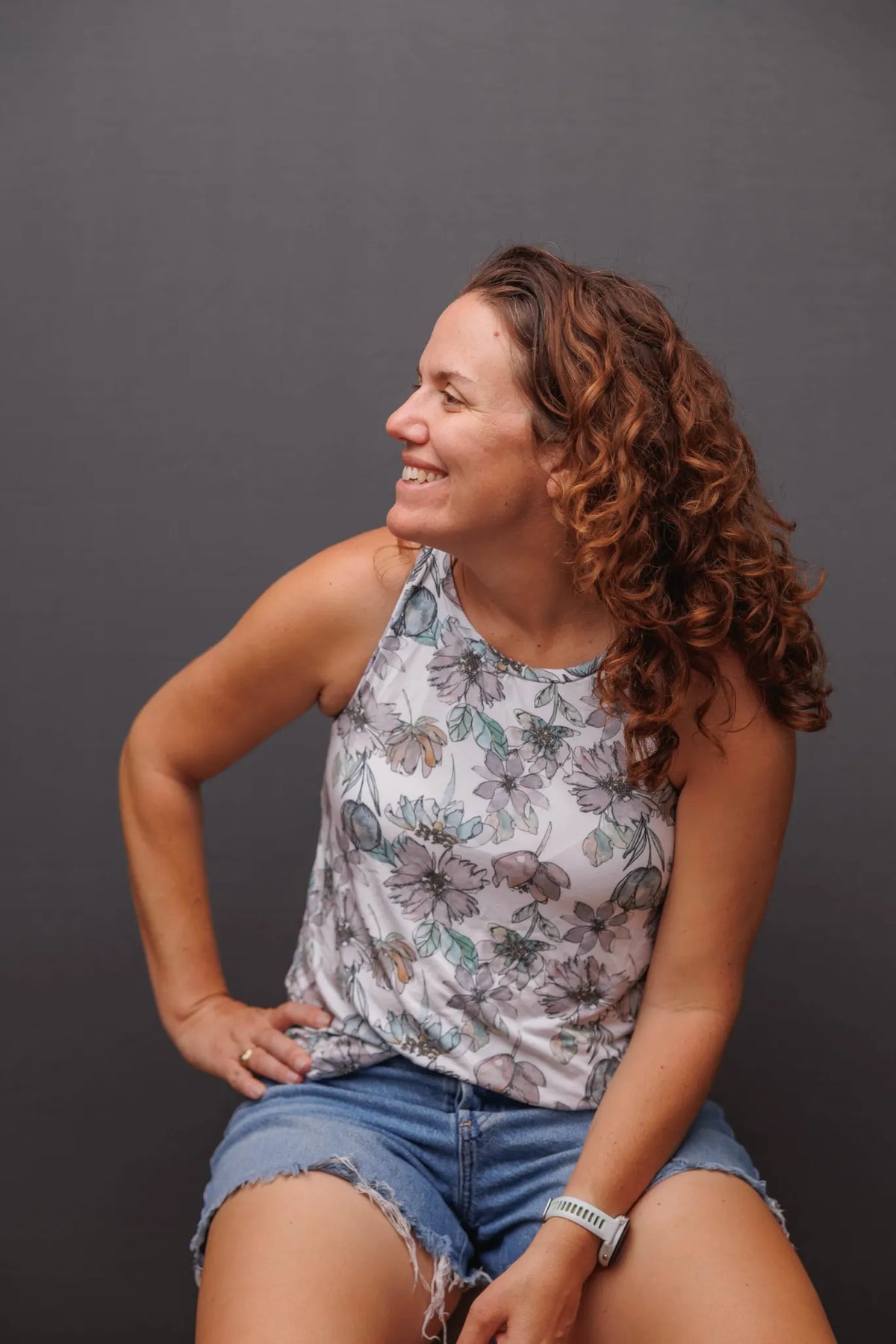 A woman with curly hair smiles, wearing a floral top and denim shorts.