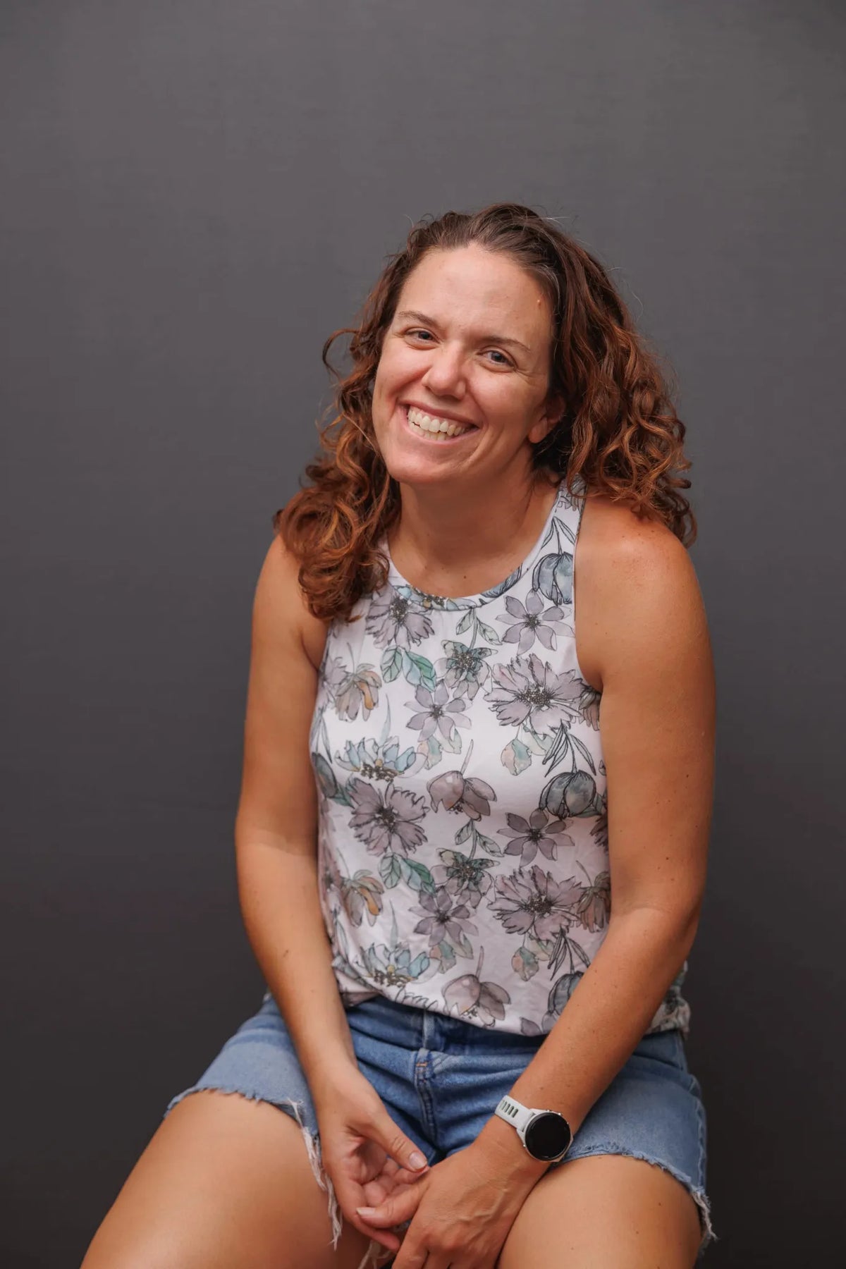 A woman with curly brown hair smiles, wearing a floral top and denim shorts.
