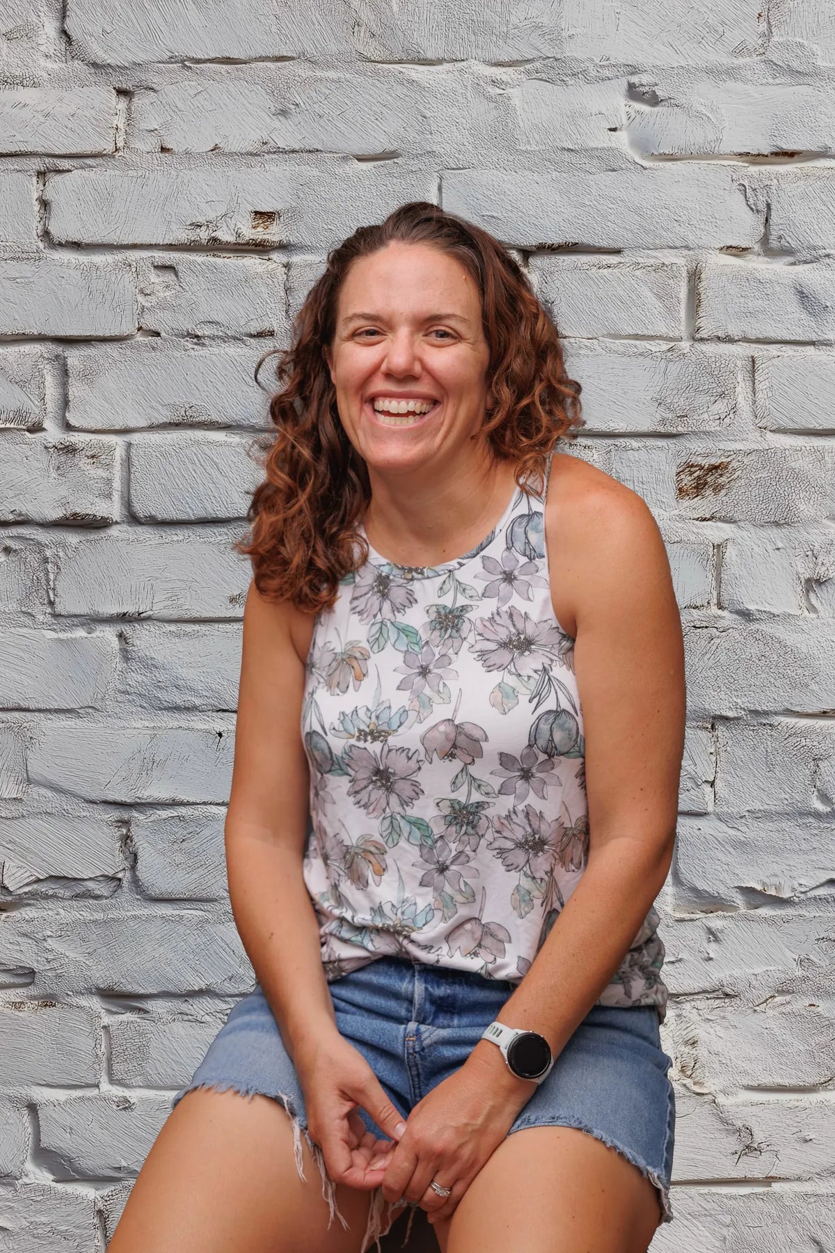 A woman with curly brown hair smiles in front of a white brick wall.