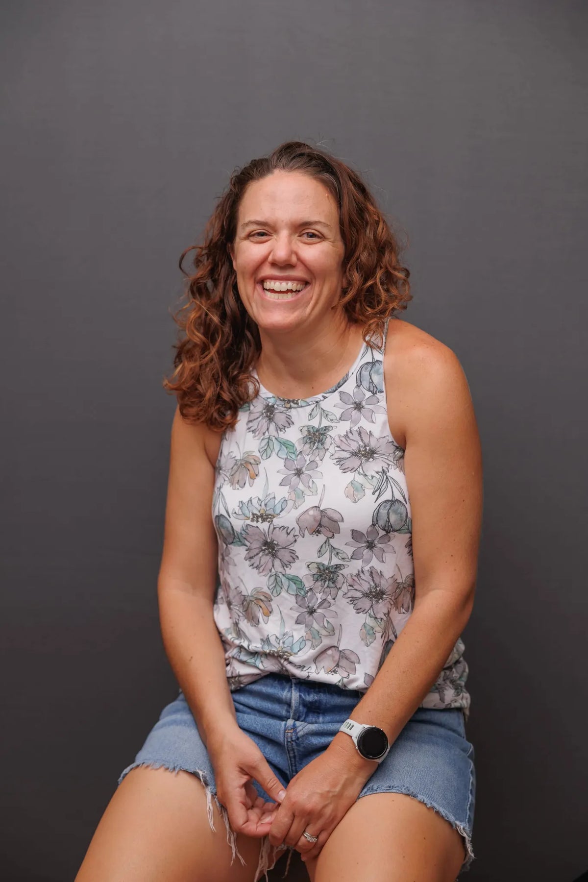 A woman with curly brown hair smiles, wearing a floral top and denim shorts.