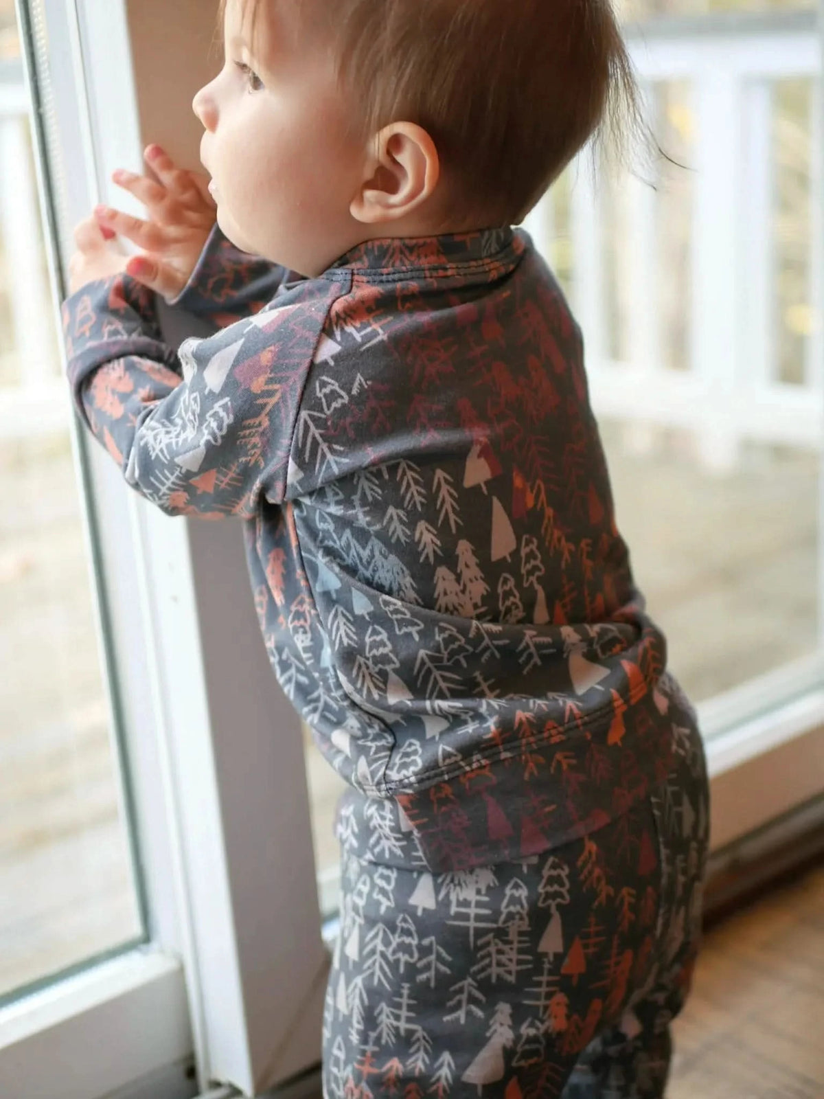 A baby in a dark fall trees patterned outfit looks out a window.