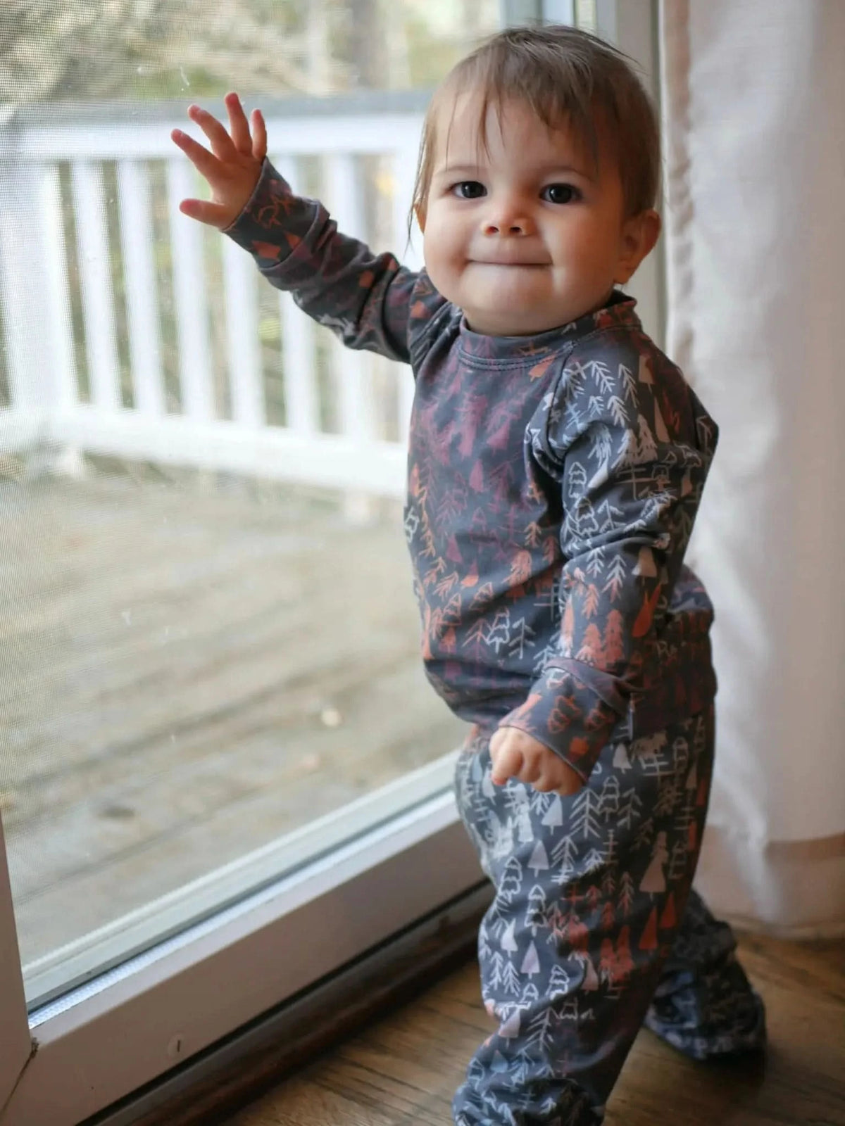 A baby in a dark fall trees patterned outfit stands by a glass door.