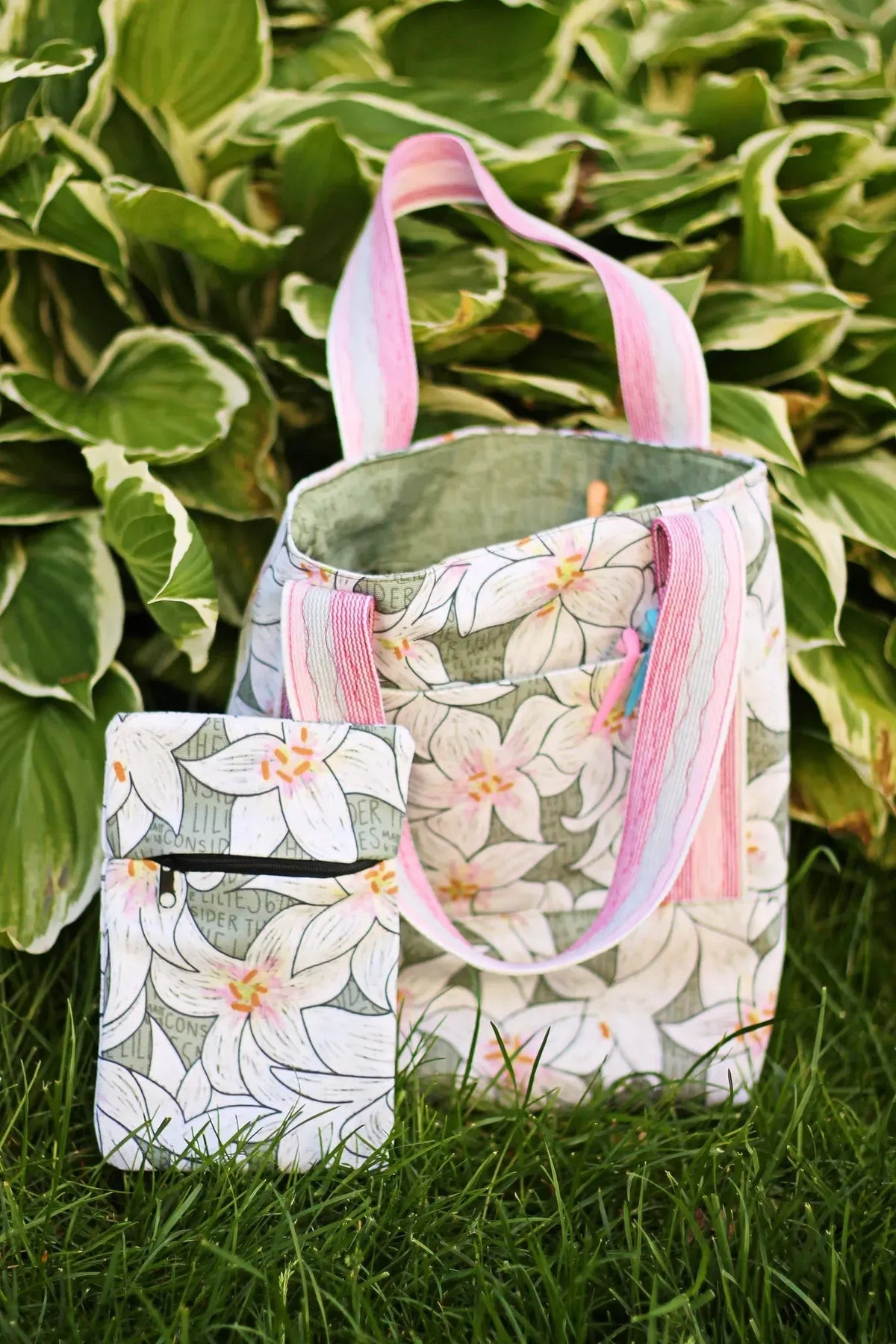A floral tote bag and matching pouch sit in grass against green foliage.