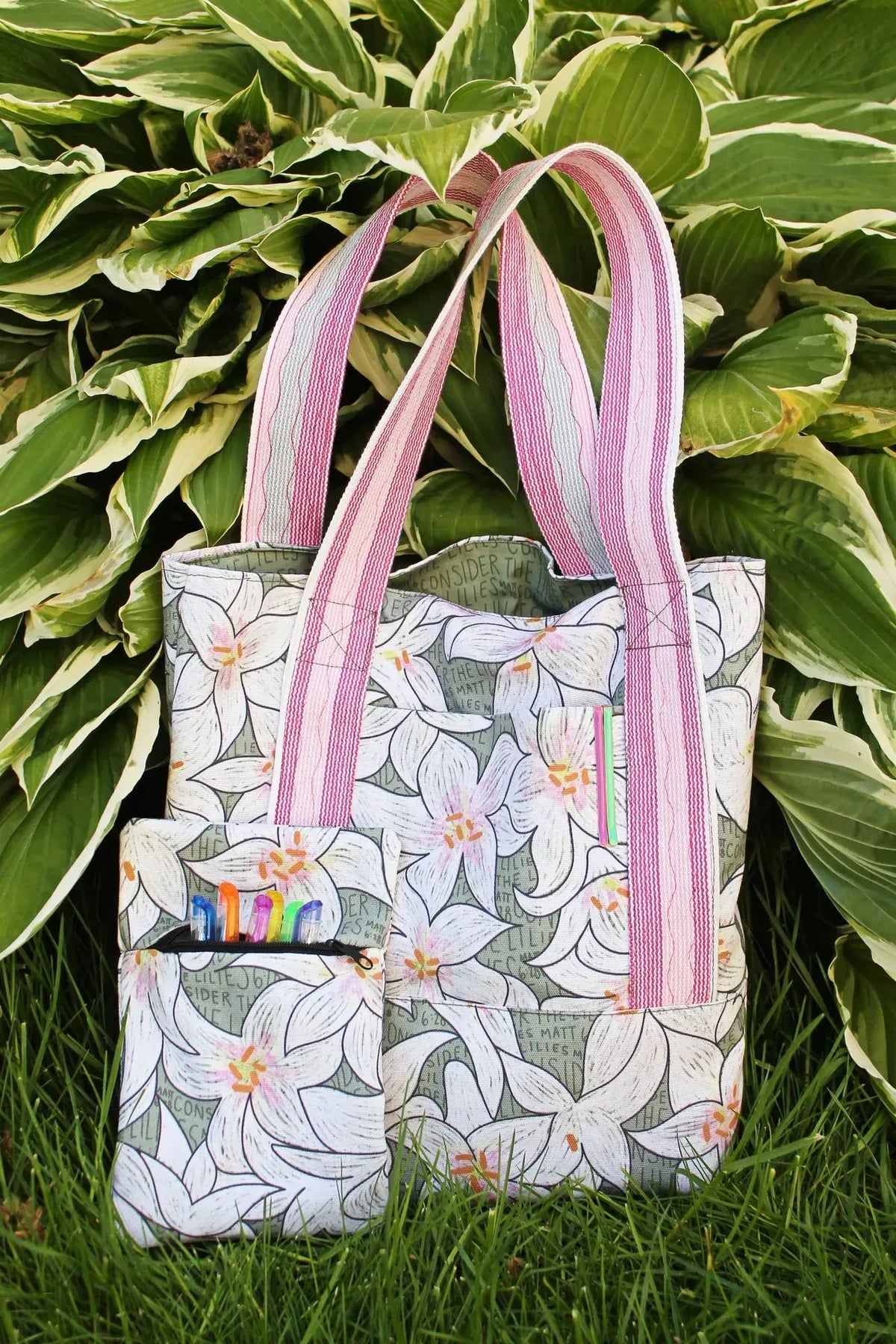 A tote bag and small pouch with a white lily pattern sit in green grass.