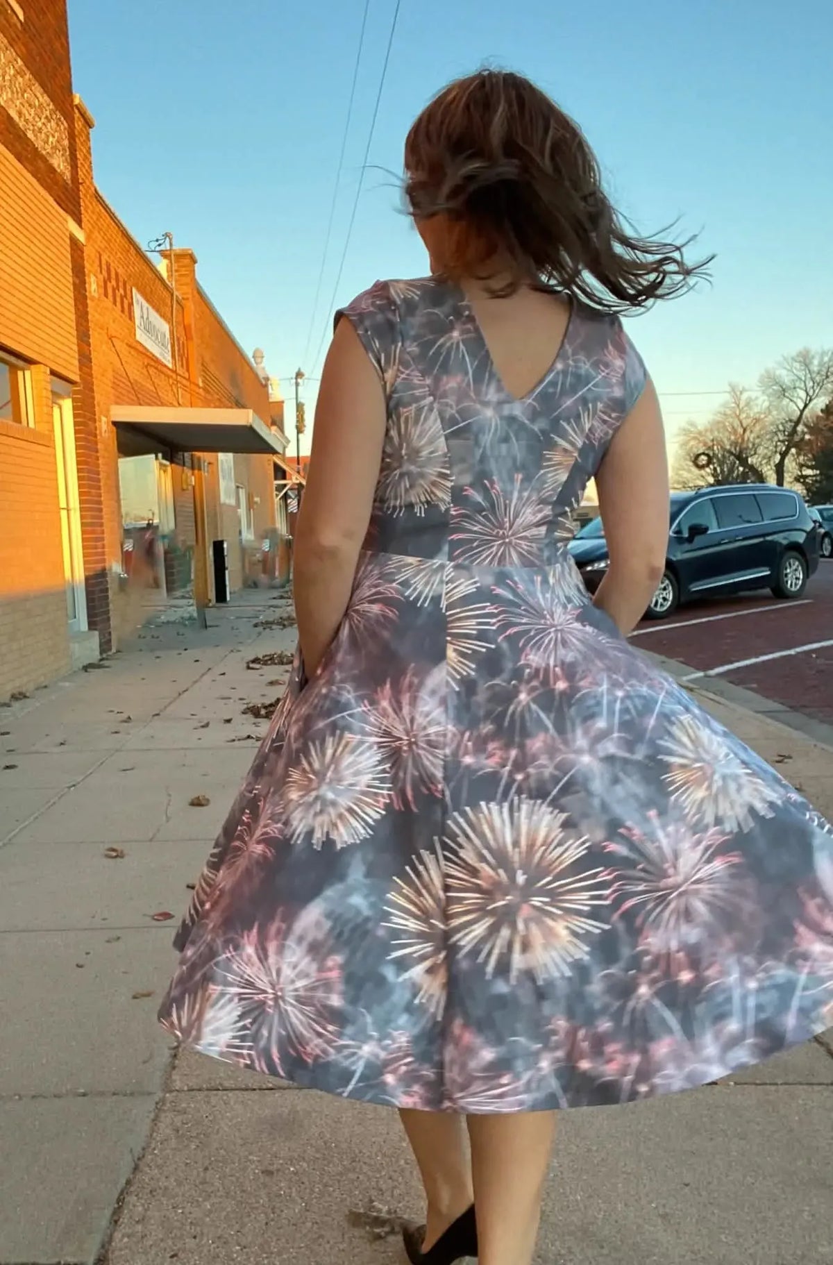 A woman in a fireworks-print dress walks away from the camera on a sidewalk.