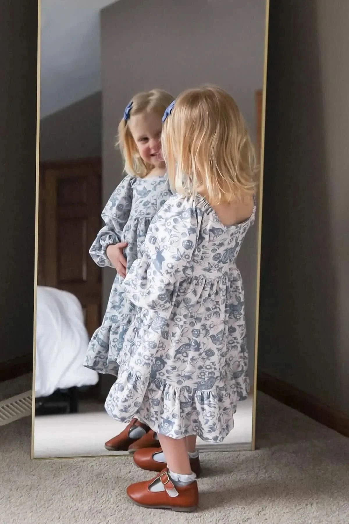 A young girl in a blue and white floral dress looks at her reflection in a mirror.
