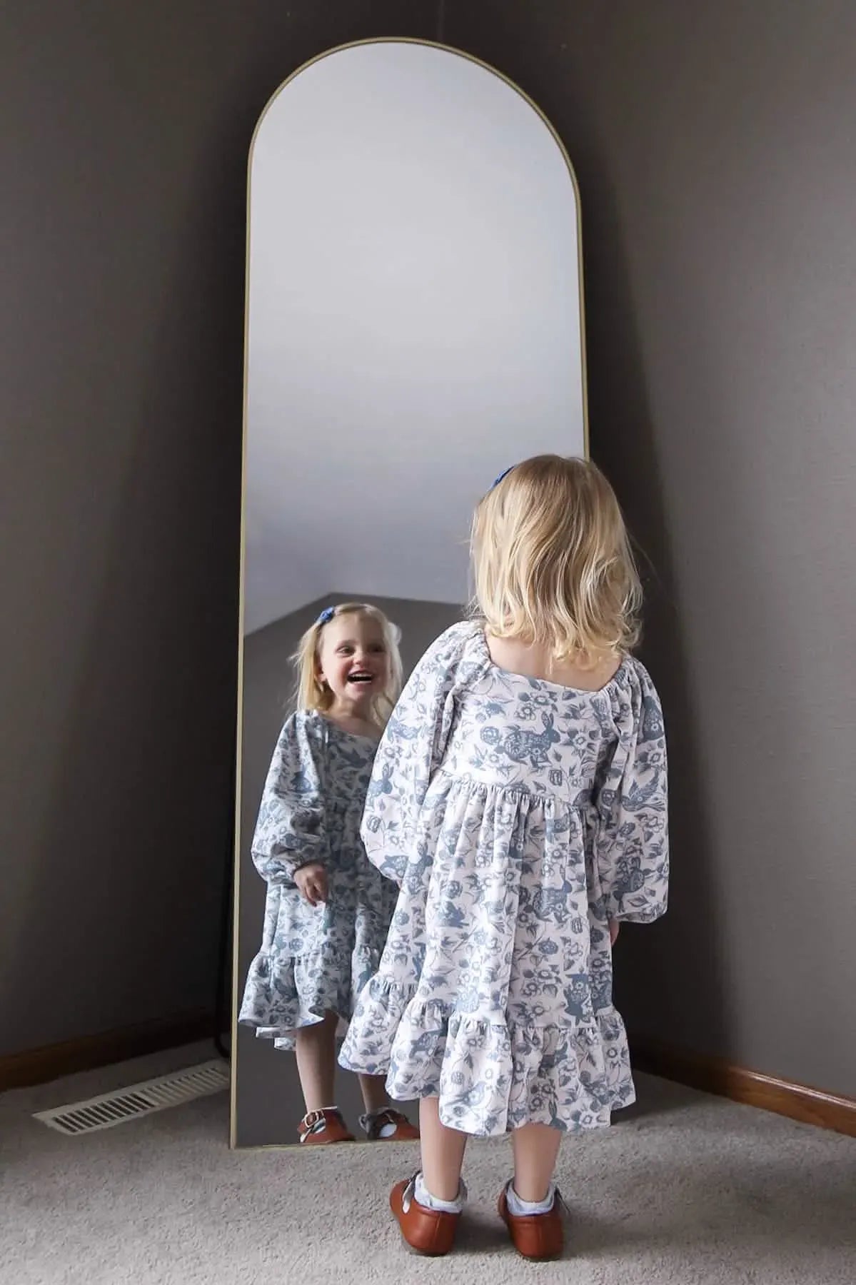 A young girl in a blue and white patterned dress looks at her reflection in a mirror.