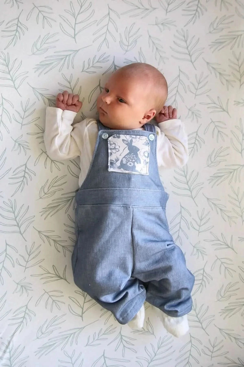 A baby in a blue linen overall with a bunny patch lies on a leaf-patterned sheet.