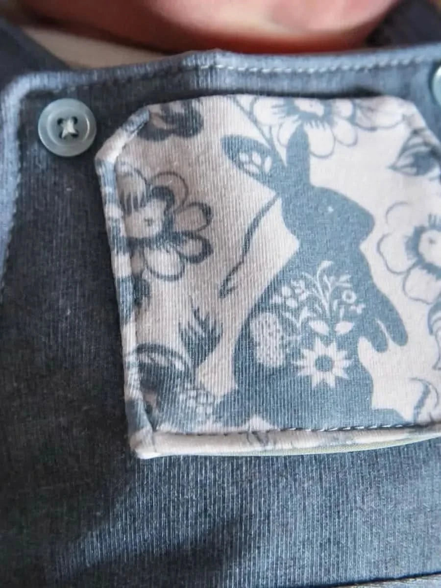 A baby wears blue overalls with a bunny and floral pattern on the chest pocket.