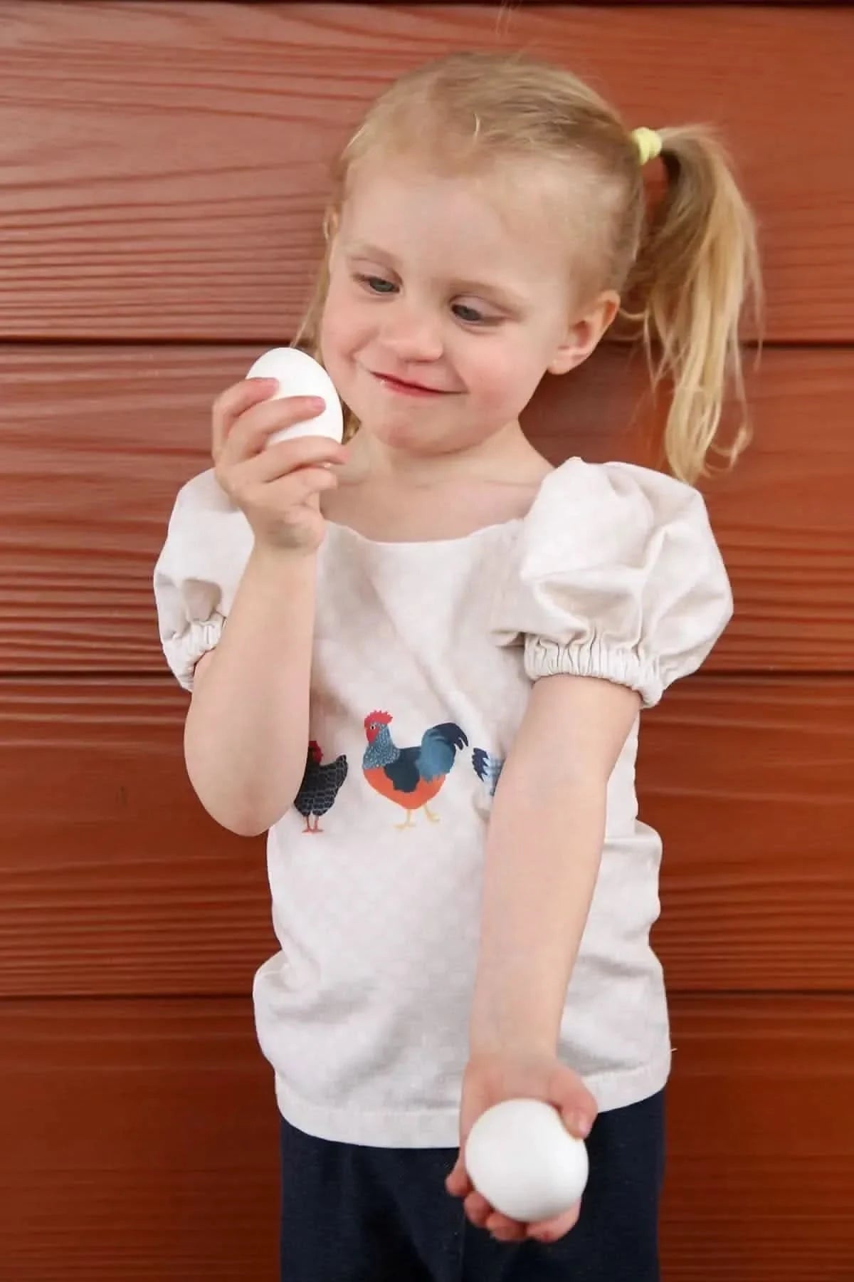 A young girl in a shirt with chicken illustrations holds two white eggs.