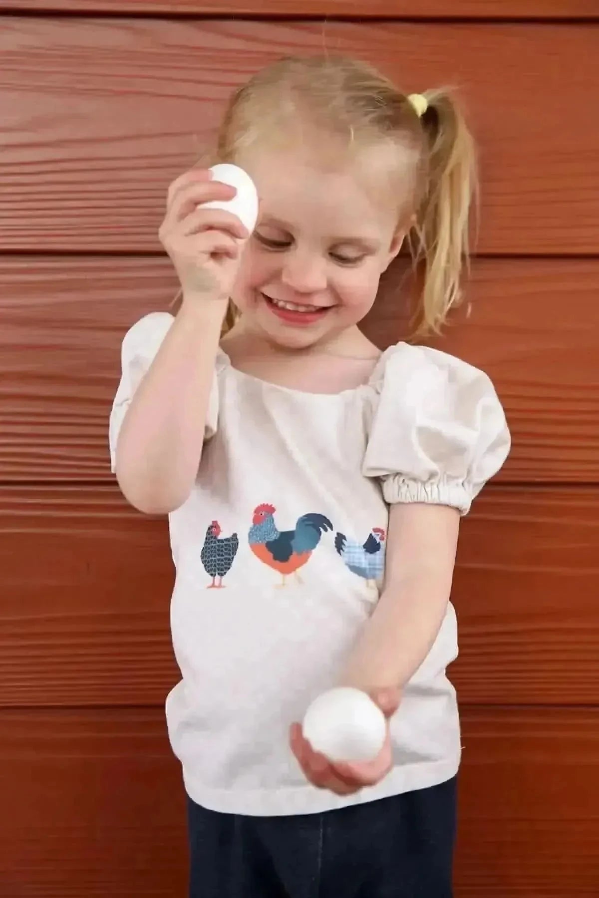 Blonde girl in chicken-print dress holding white eggs against wooden wall.
