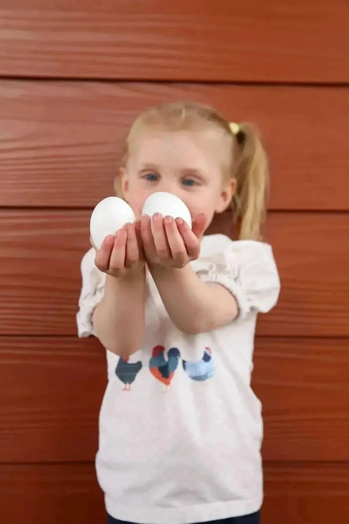 Girl in chicken-print dress holding two white eggs against wooden wall.
