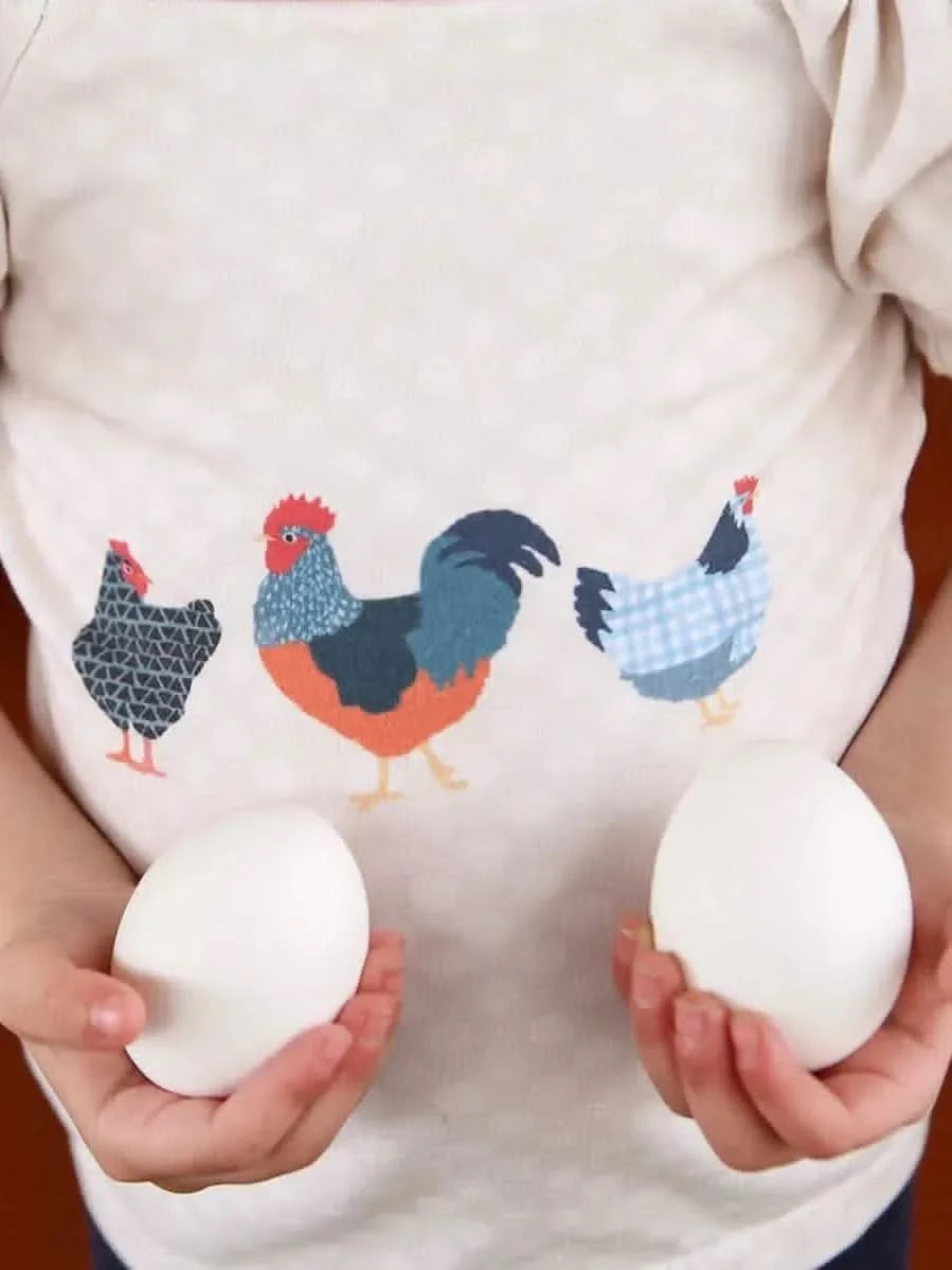 A child in a chicken-print shirt holds two white eggs.