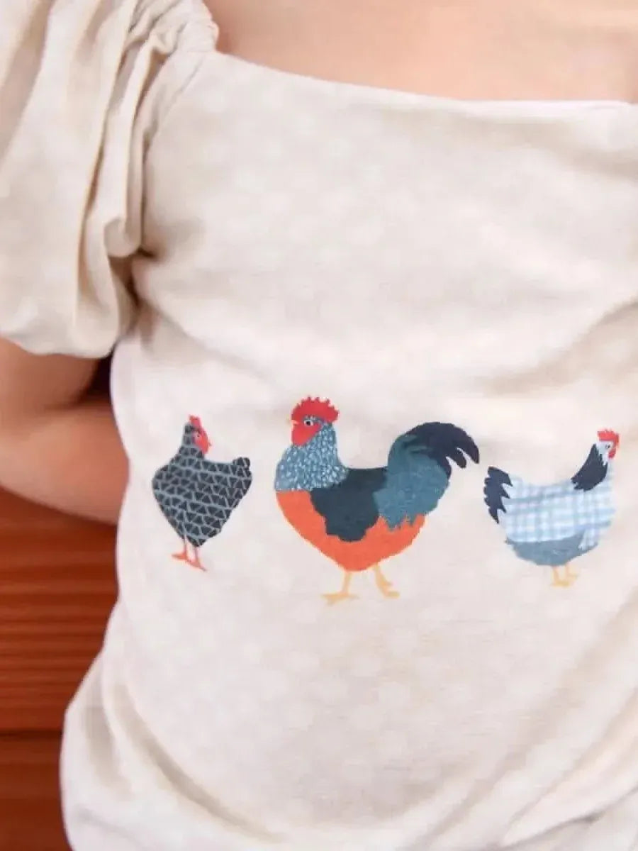 A child wears a light-colored shirt with three colorful patchwork chickens printed on the front.