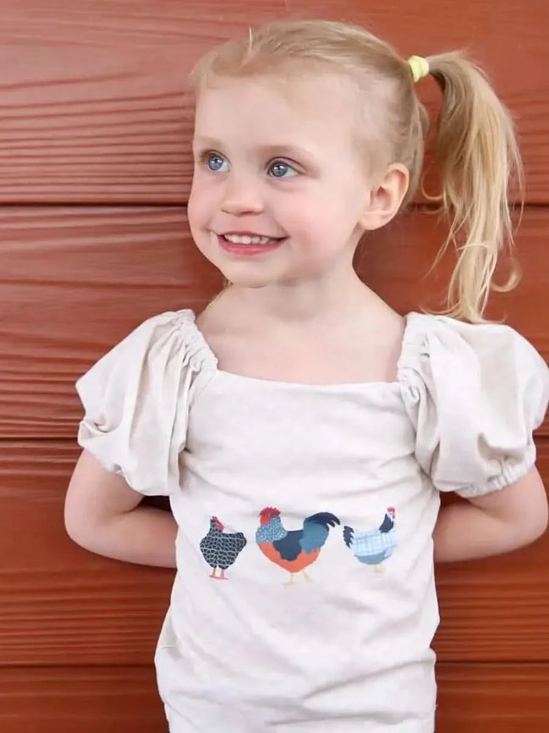 A young girl smiles, wearing a light-colored top with three patchwork chickens.