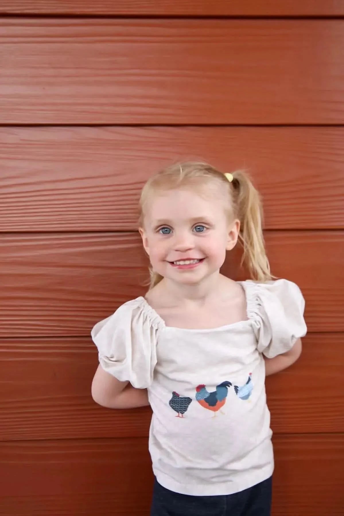 A young girl smiles, wearing a shirt with three patchwork chickens.