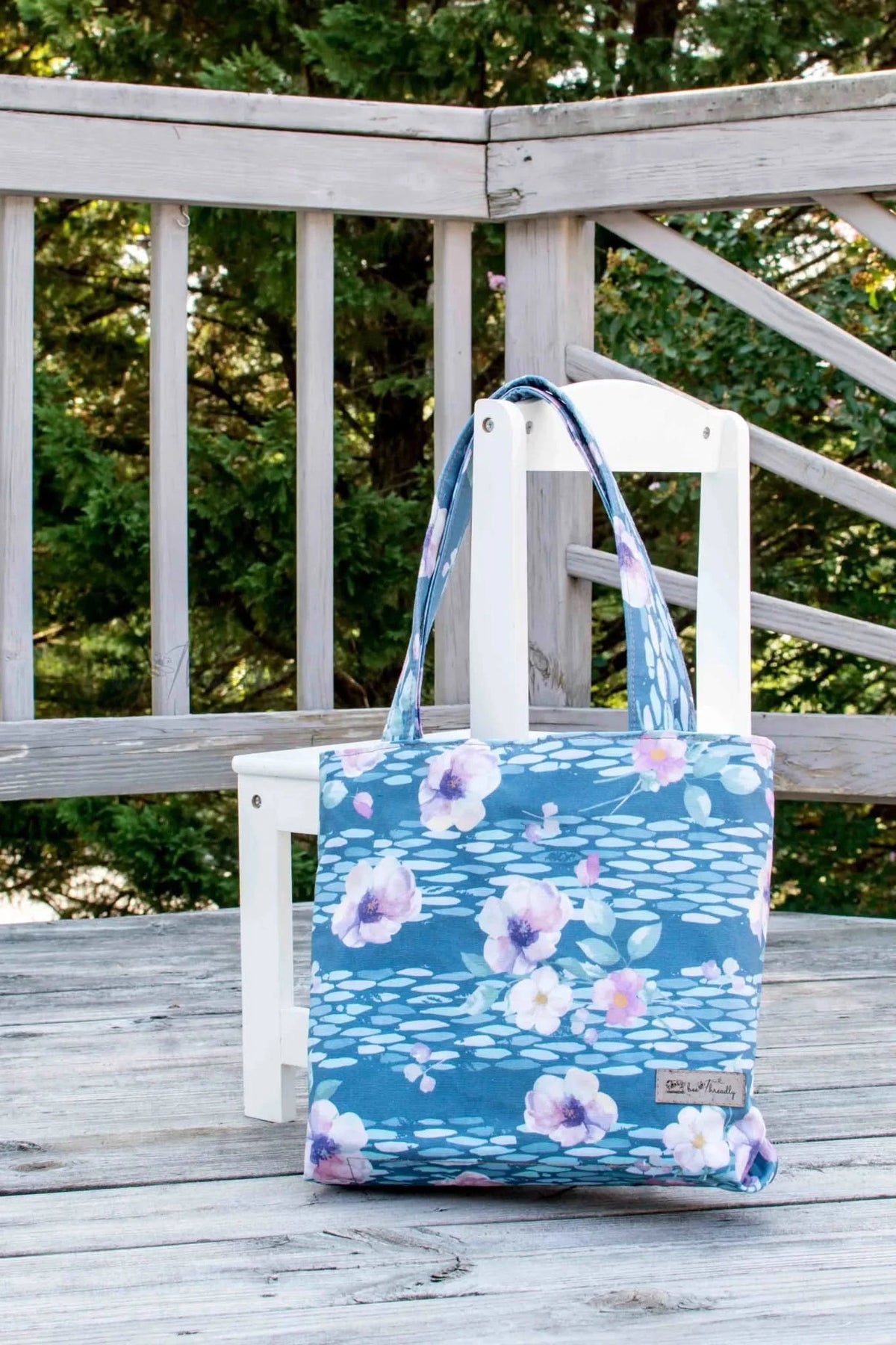 A blue tote bag with pink and white watercolor anemones sits on a wooden deck.