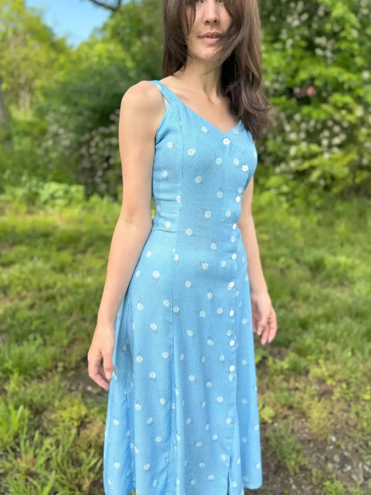 A woman in a blue polka dot dress with white buttons stands in a grassy area.