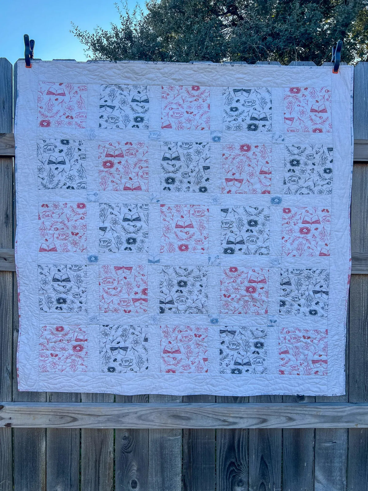 A quilt with red and black toile patterns hangs on a wooden fence.
