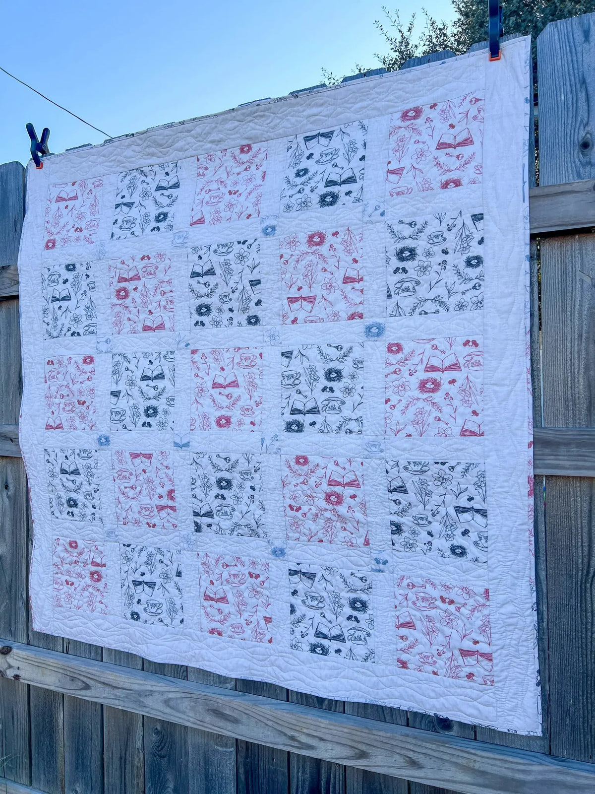 A white quilt with red and blue toile patterns hangs on a wooden fence.