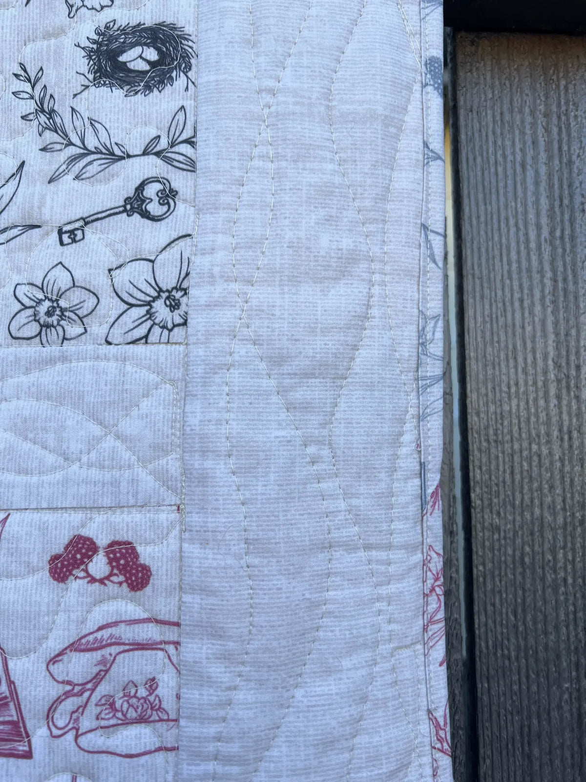 A quilted fabric with black and pink toile patterns, next to a dark wooden surface.