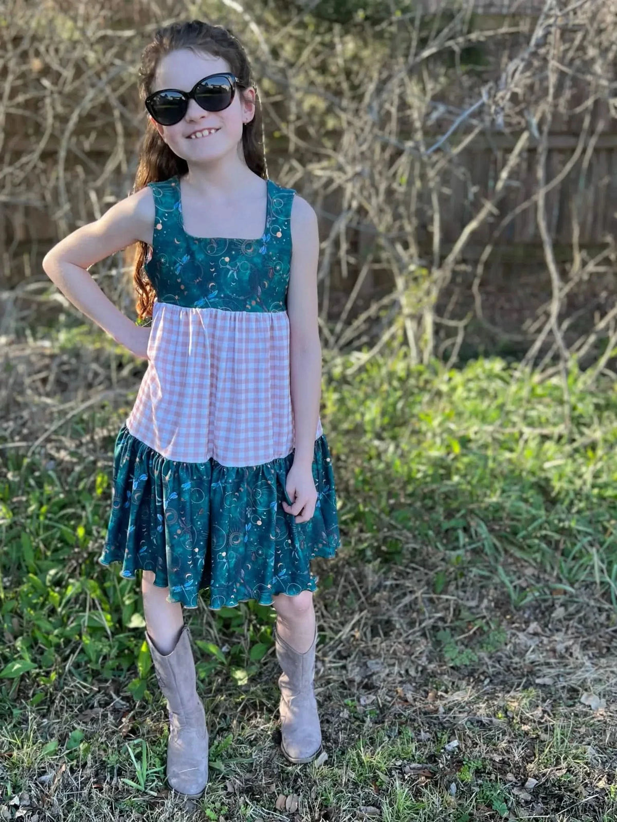 A young girl in a tan gingham and teal dragonfly dress with cowboy boots.
