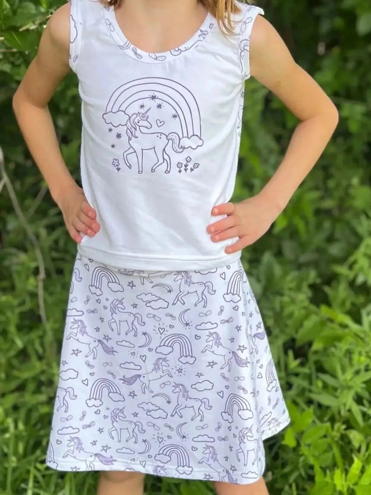 Young girl wearing a white tank top with unicorn and rainbow outline and a purple skirt with unicorn print.