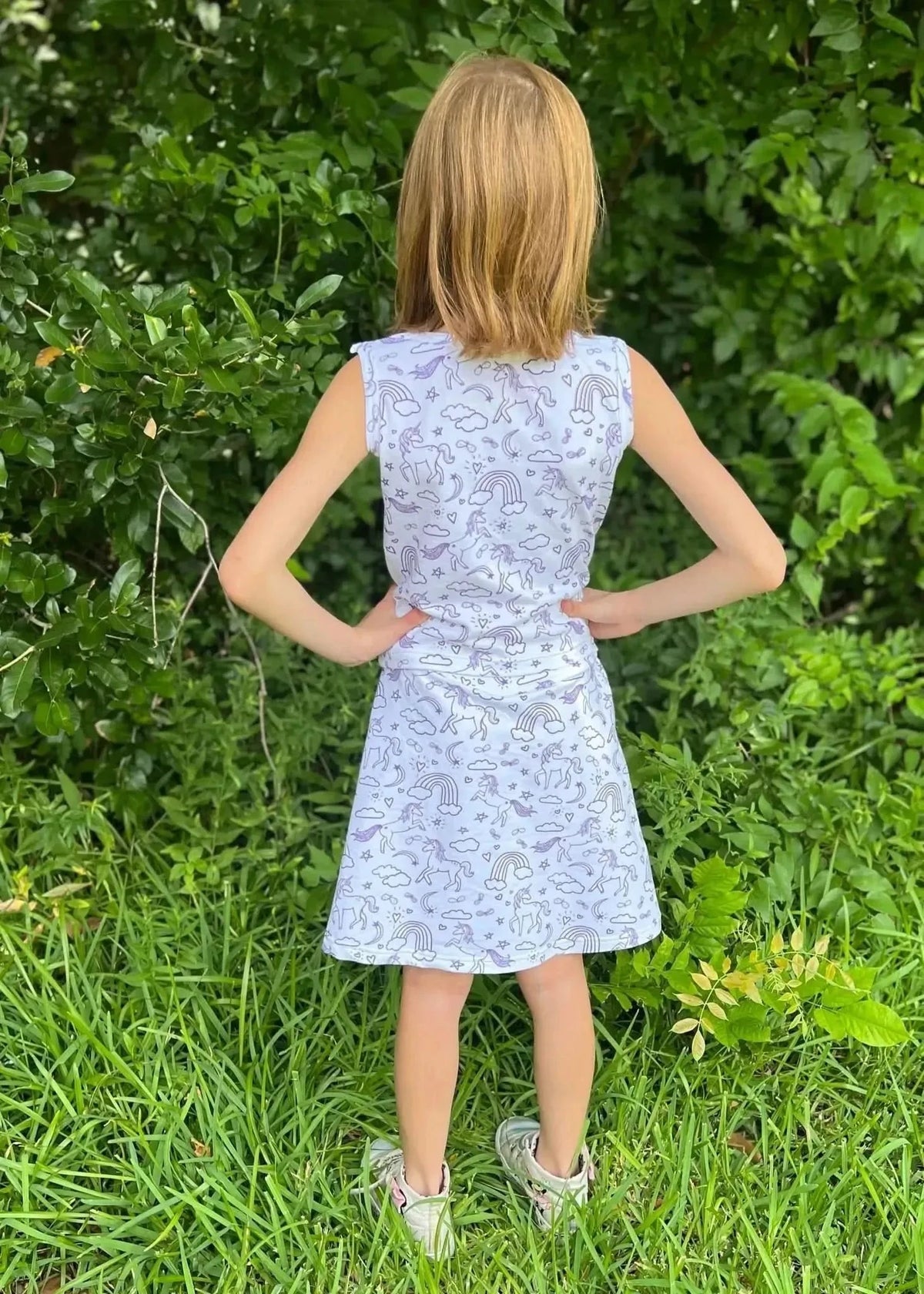 Rear view of a girl wearing a sleeveless white dress with unicorn print, standing in grass.