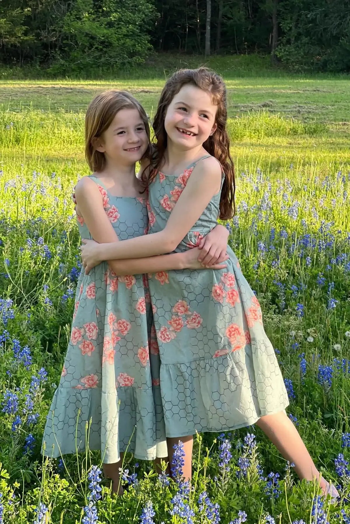 Two girls in matching honeycomb floral dresses stand in a field of bluebonnets.