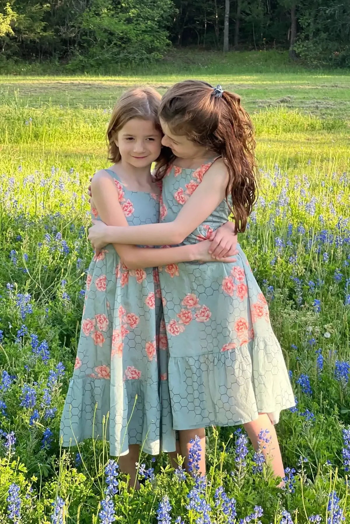 Two girls in matching honeycomb floral dresses stand in a field of bluebonnets.