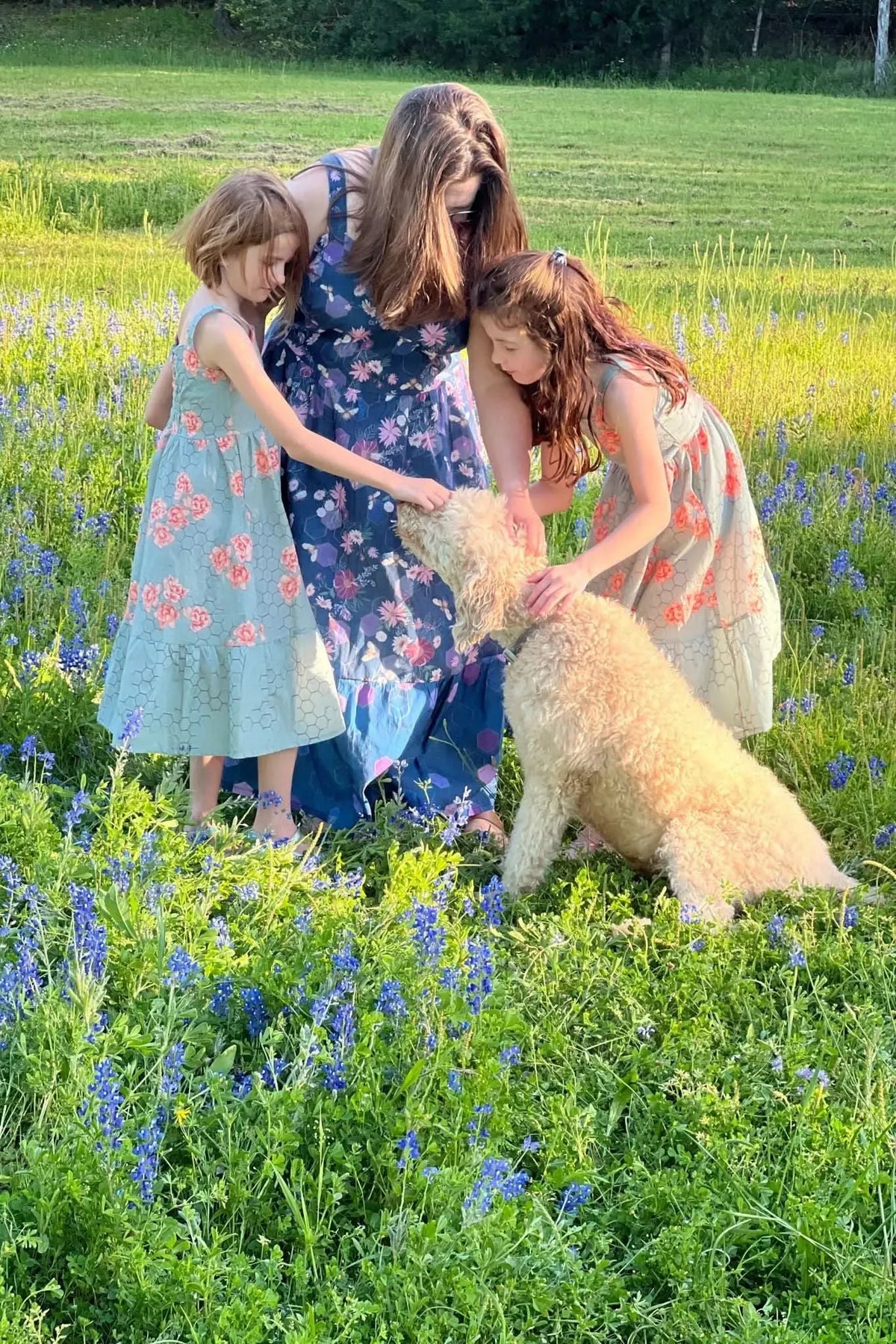 A woman and two girls in dresses pet a dog in a field of blue flowers.