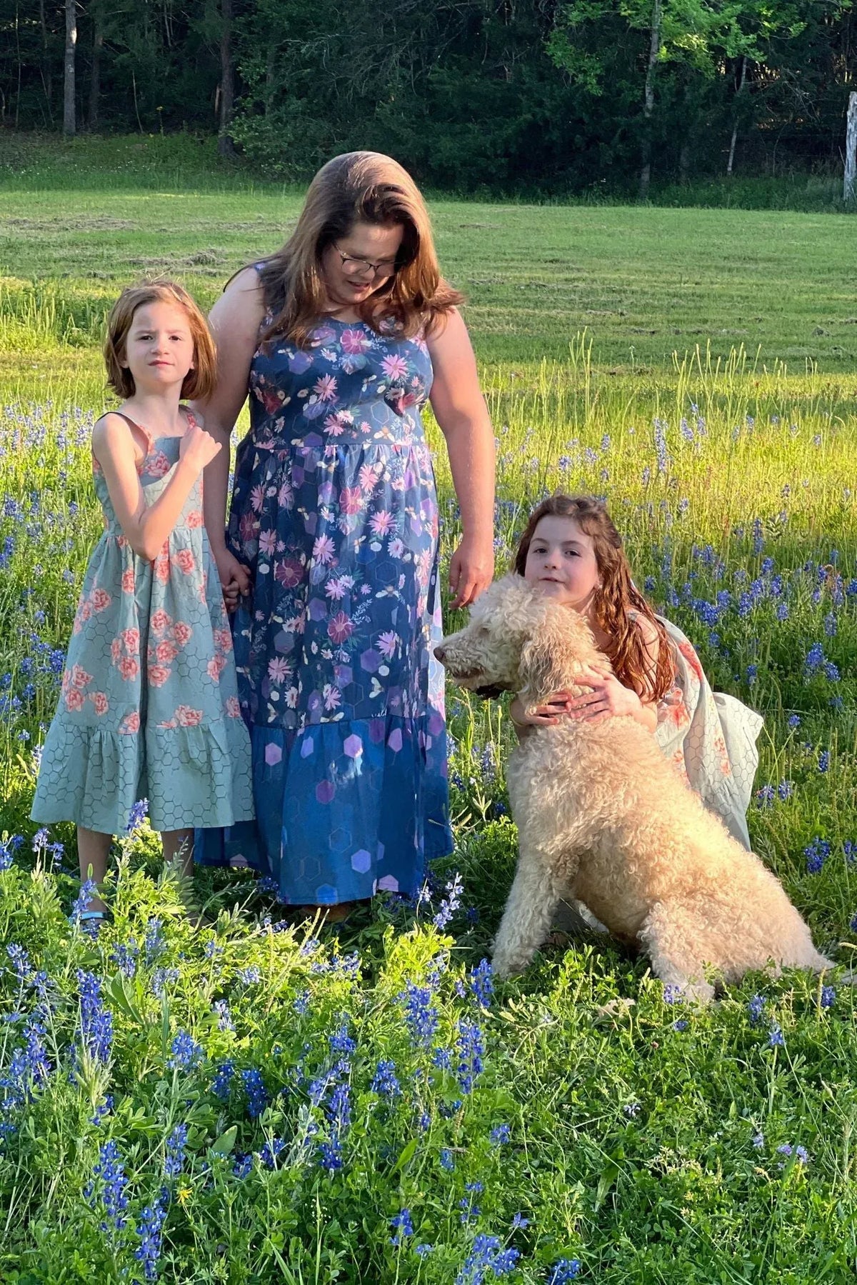 A woman and two girls in floral dresses stand in a field of bluebonnets with a dog.