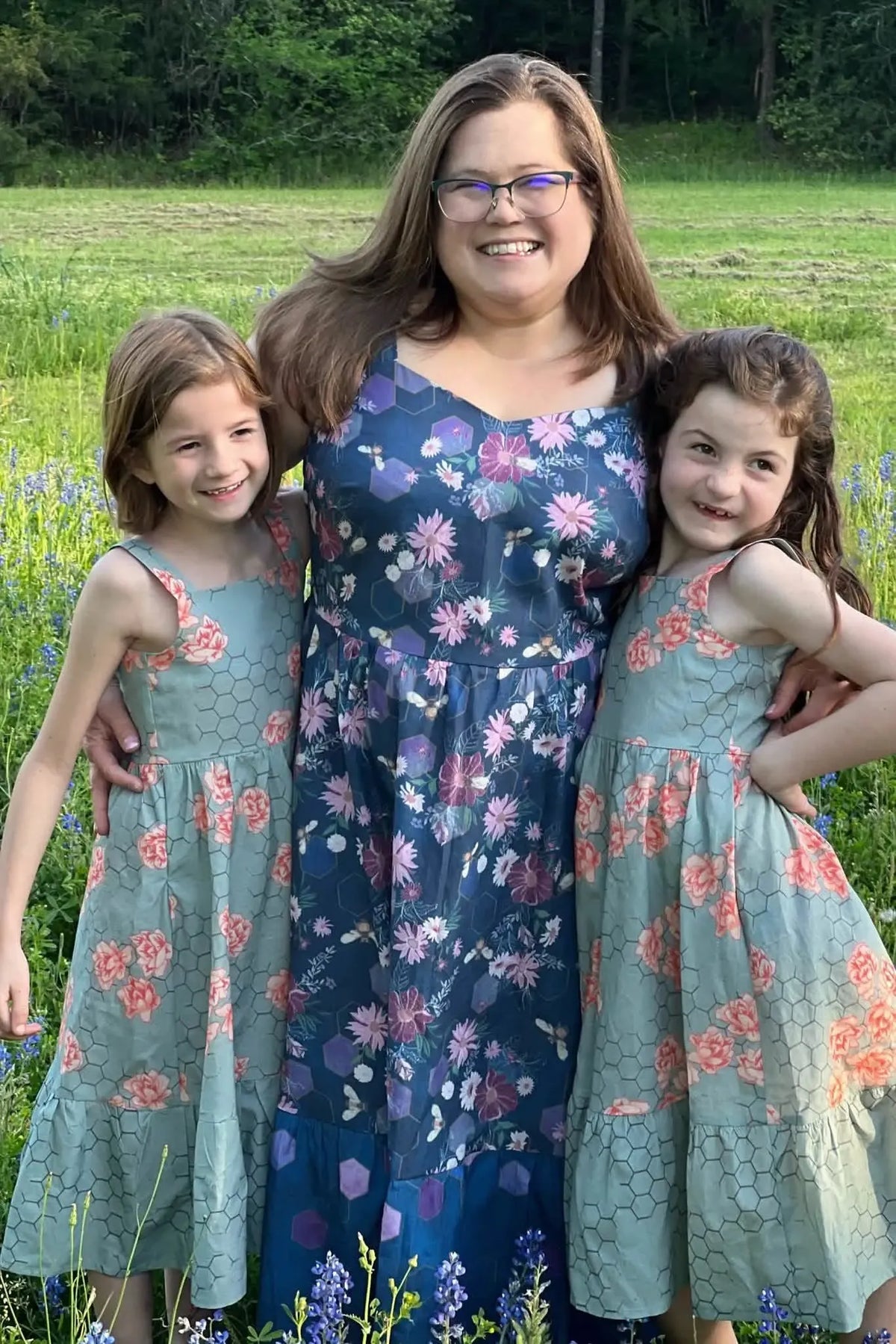 A woman and two girls wear dresses with a purple floral and bee pattern.