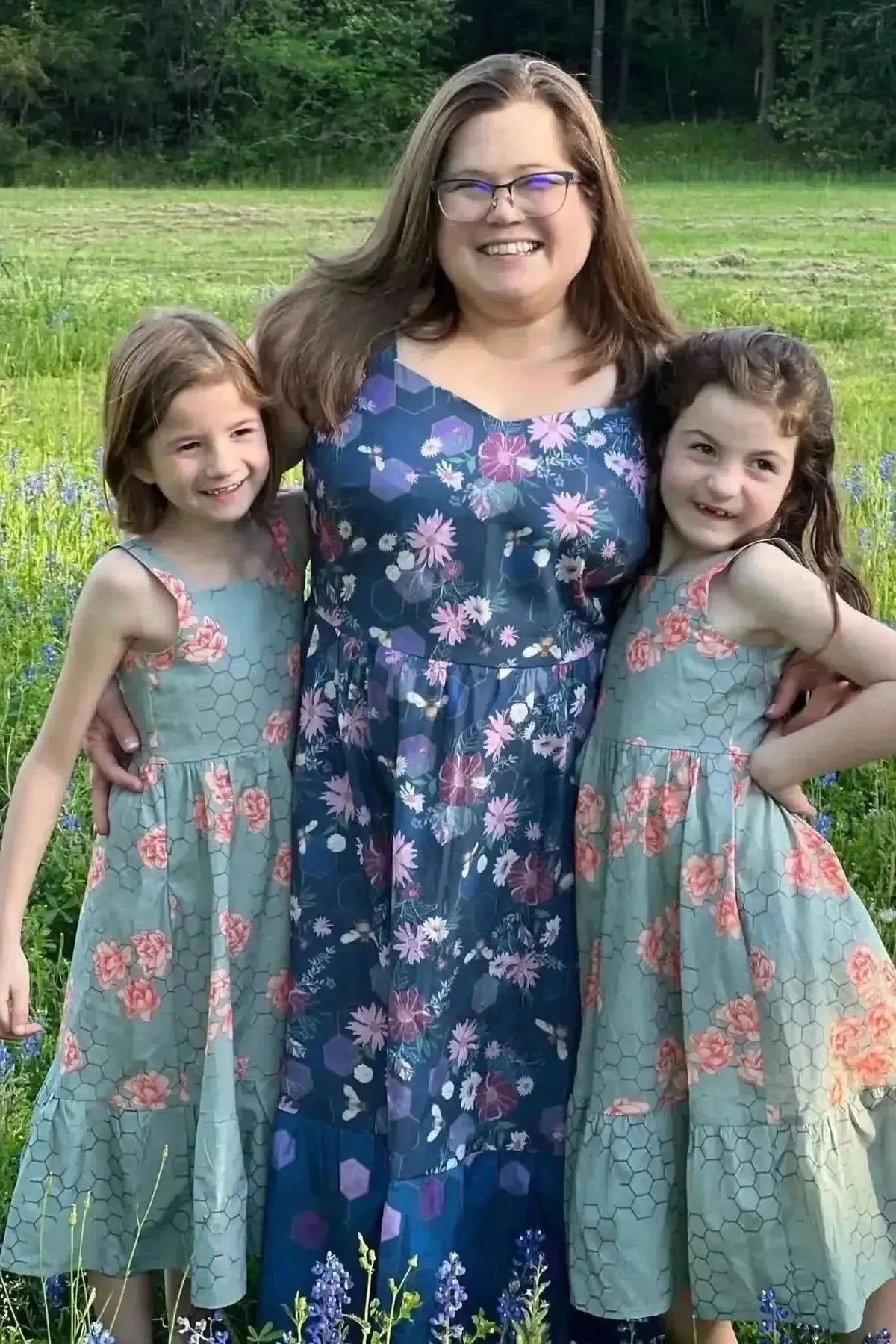 Purple fabric with garden bees print, shown in matching dresses on a woman and two girls outdoors.