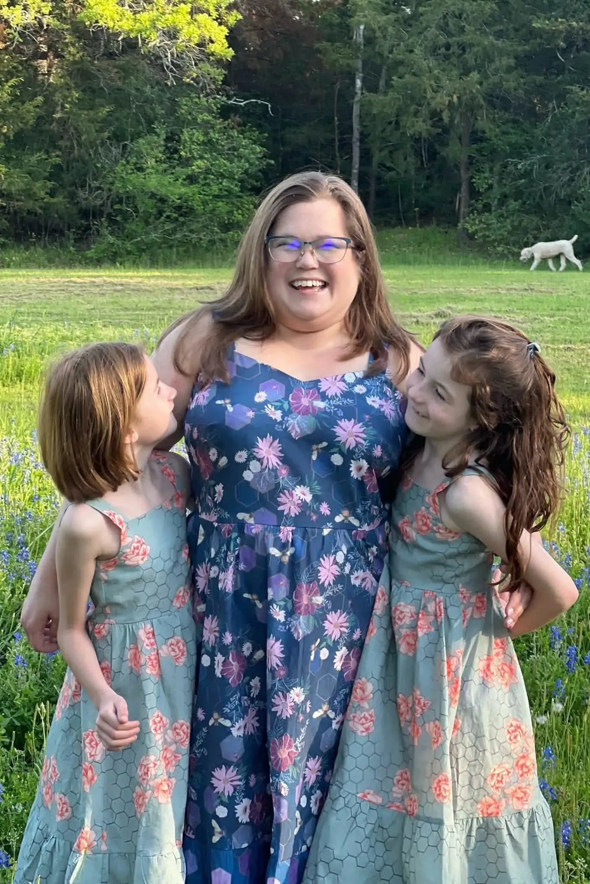 A woman and two girls in floral dresses stand in a field with a dog in the background.
