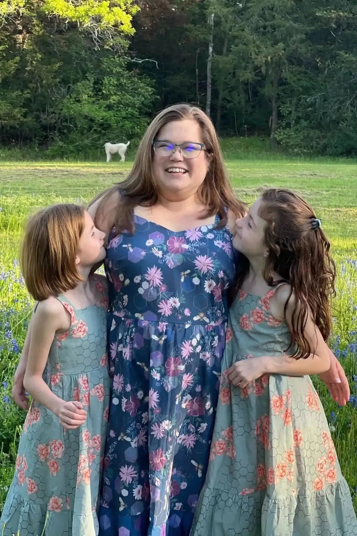 Purple fabric with garden bees print, shown in matching dresses on a woman and two girls outdoors.