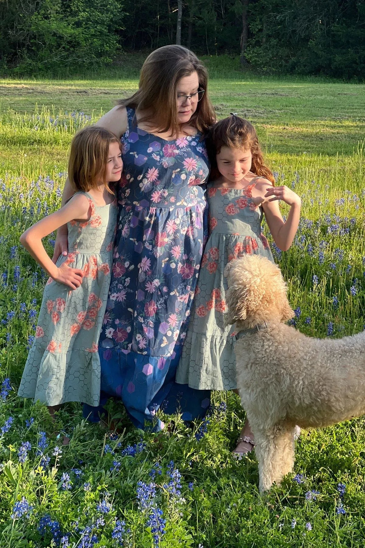A woman and two girls wear dresses with a purple floral and bee pattern.