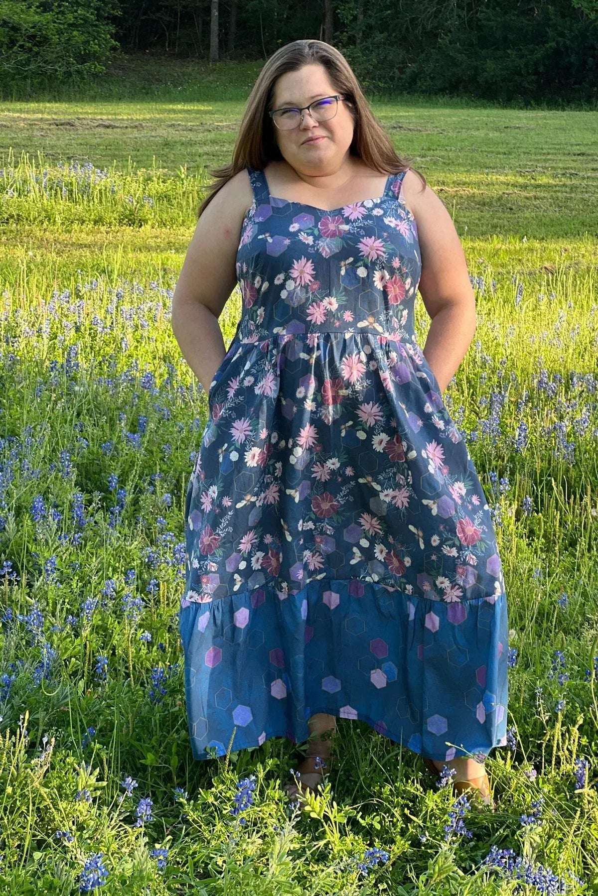 A woman in a floral dress with purple hexagons stands in a field of blue flowers.