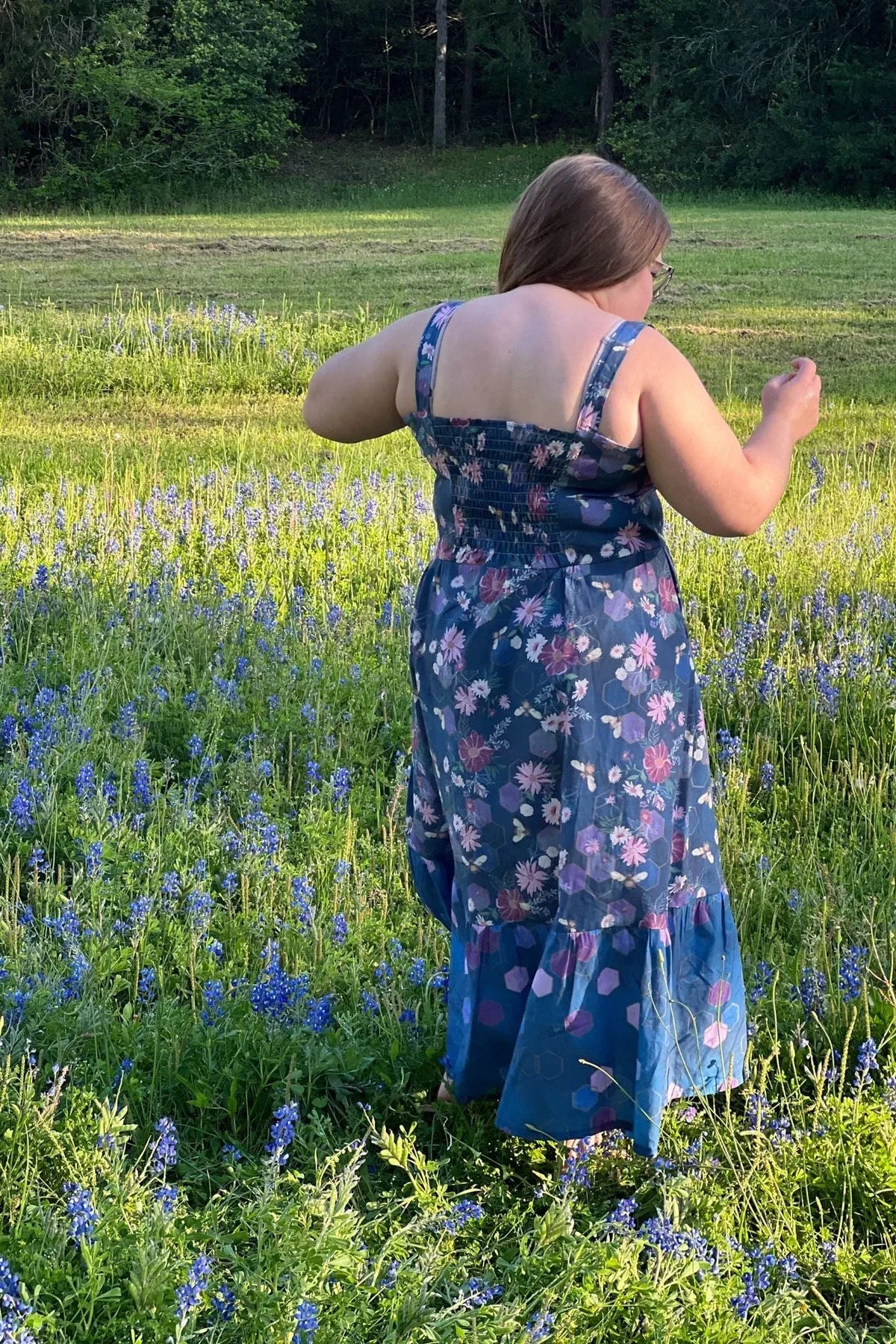 A person from behind in a floral dress with bees and purple hexagons in a field of blue flowers.