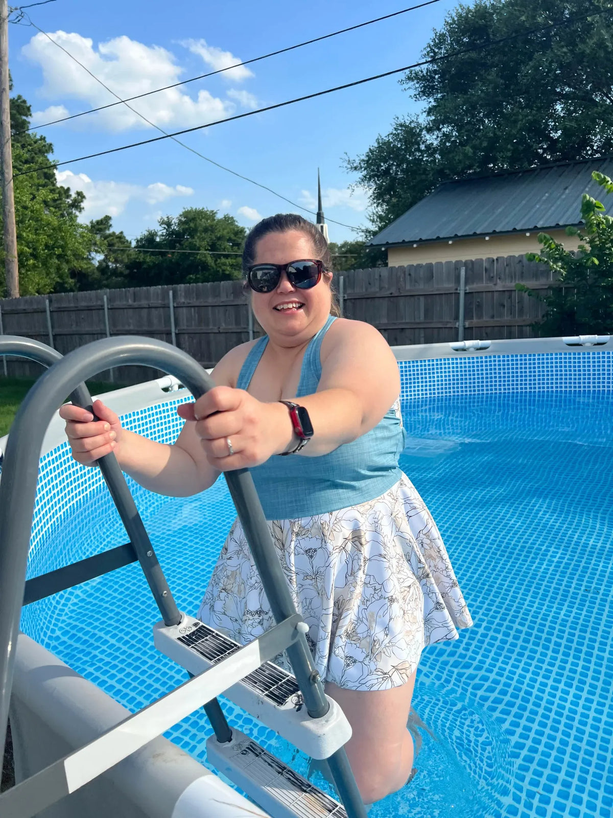 A woman in a blue top and floral skirt stands on pool steps, smiling.