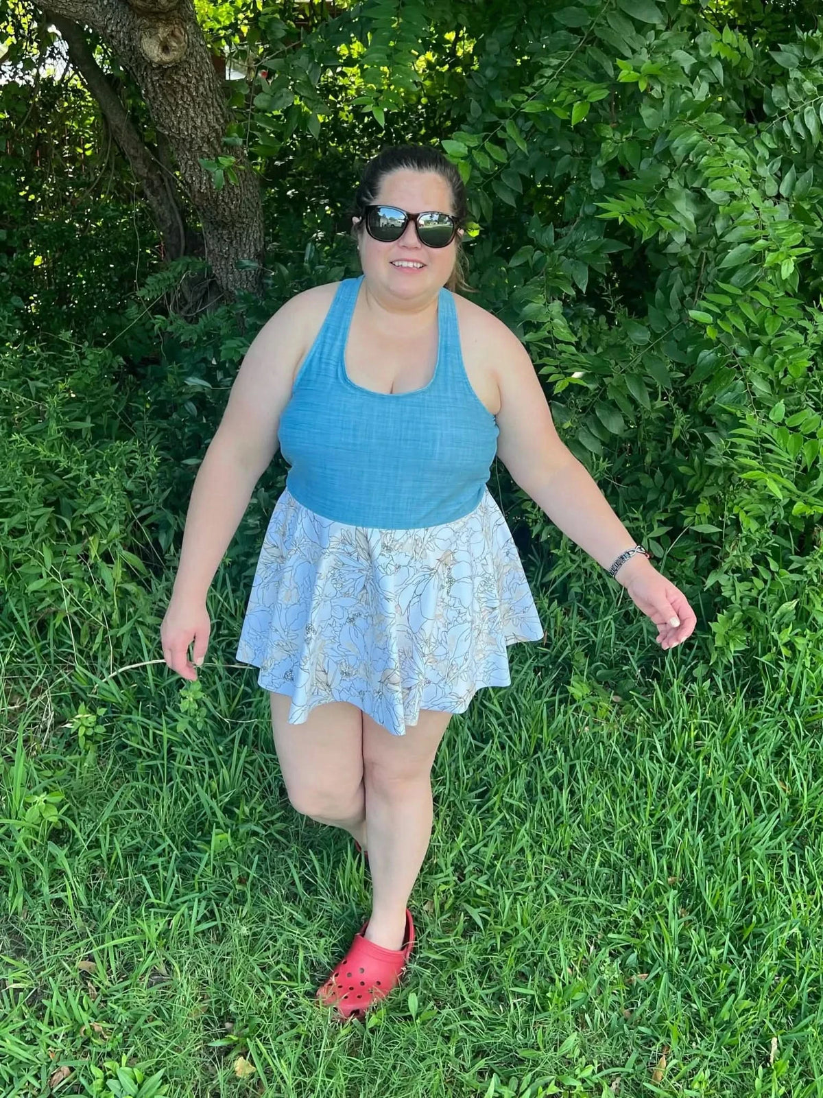 A woman in a blue tank top and floral skirt stands in grass.