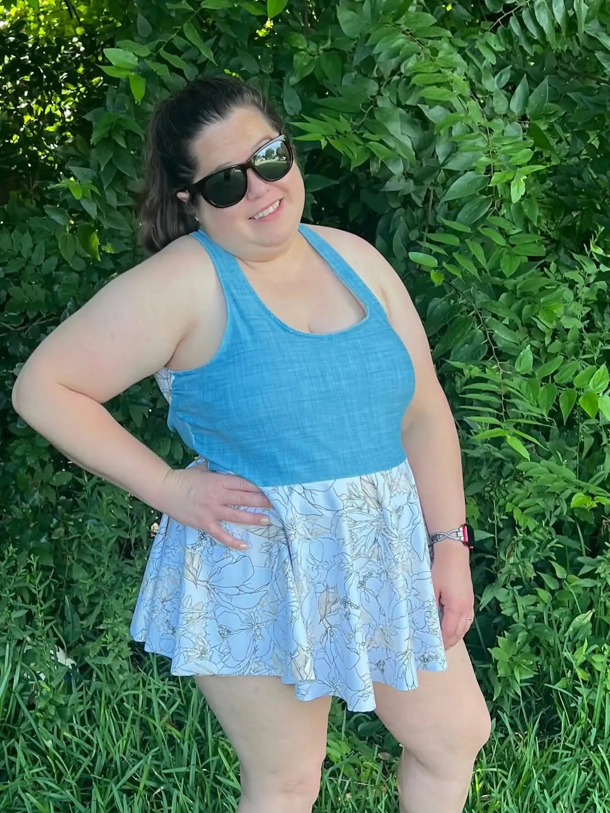 A woman in a blue top and floral skirt stands smiling in front of green foliage.