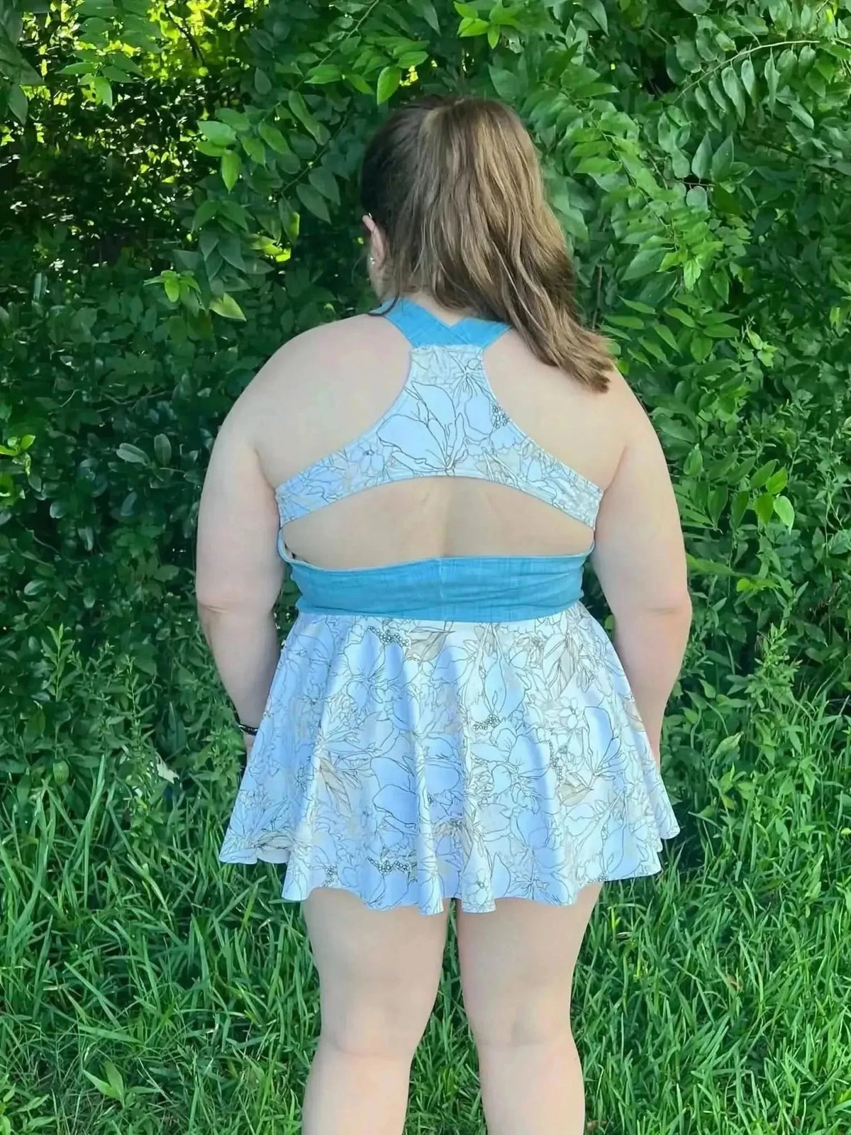 Rear view of a girl in a light blue botanical print skater dress with racerback straps, against green foliage.