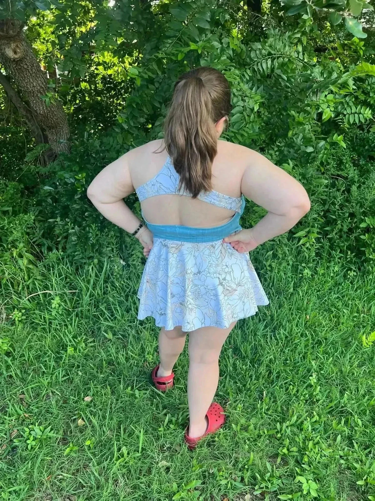 Rear view of a woman in a light blue backless swimsuit with floral botanical print, in a green outdoor setting.