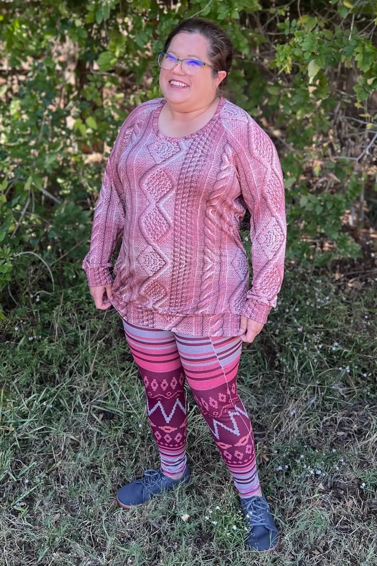 A woman in a rose cable knit top and patterned leggings smiles outdoors.