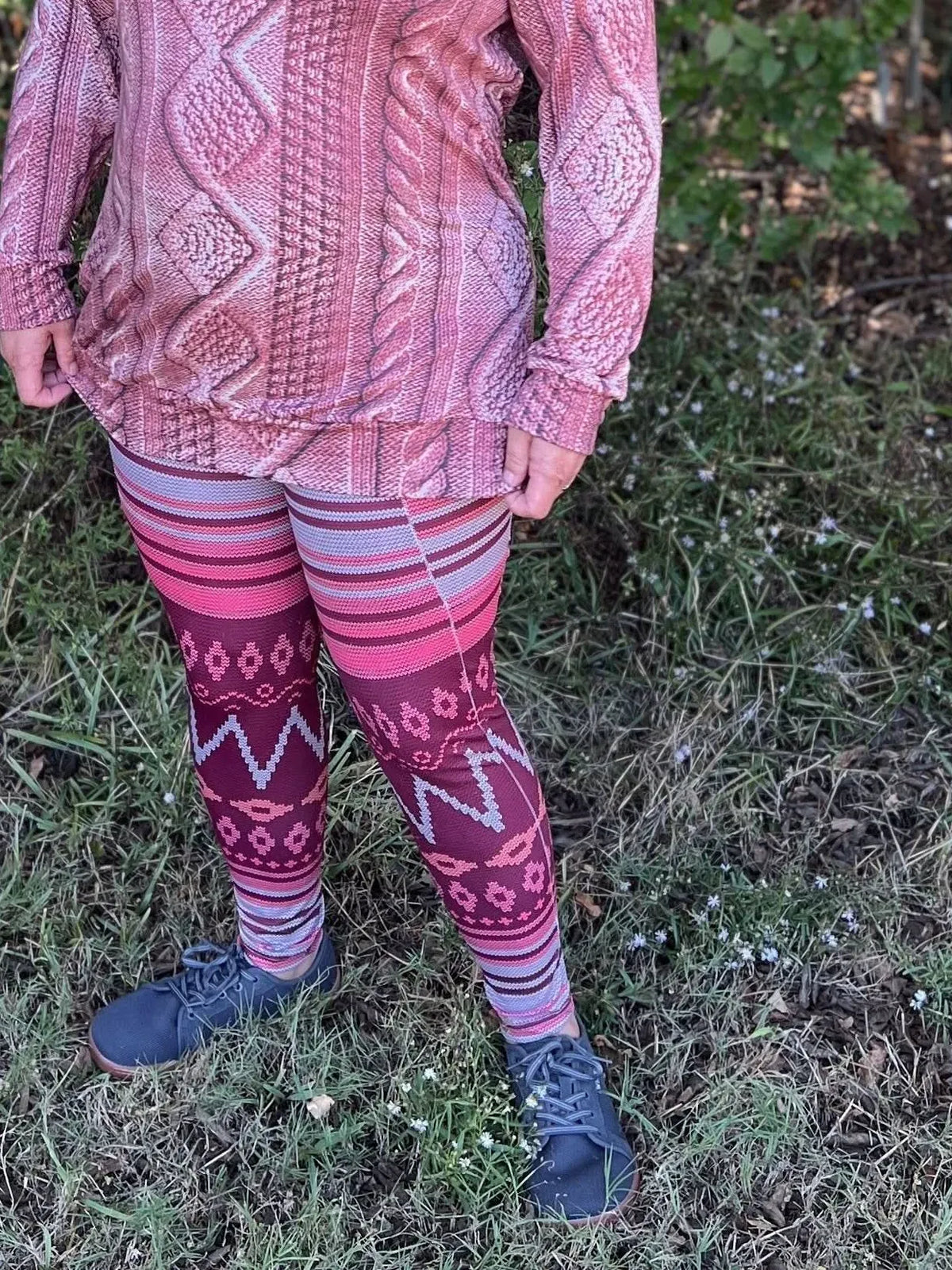 A person wearing a pink cable knit top and patterned leggings stands on grass.