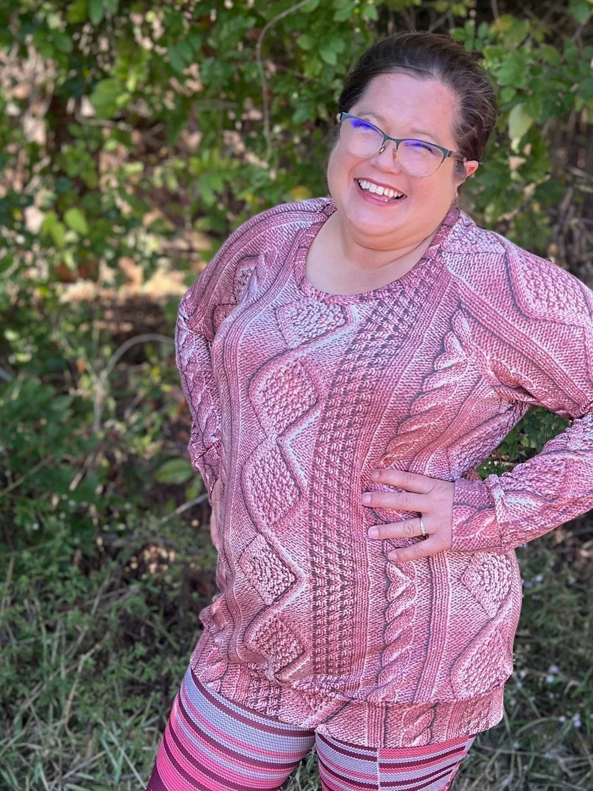 A woman smiles, wearing a rose cable knit top and striped leggings.