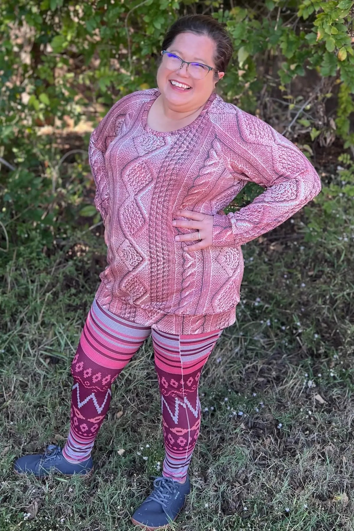 A woman smiles, wearing a pink cable knit top and patterned leggings.