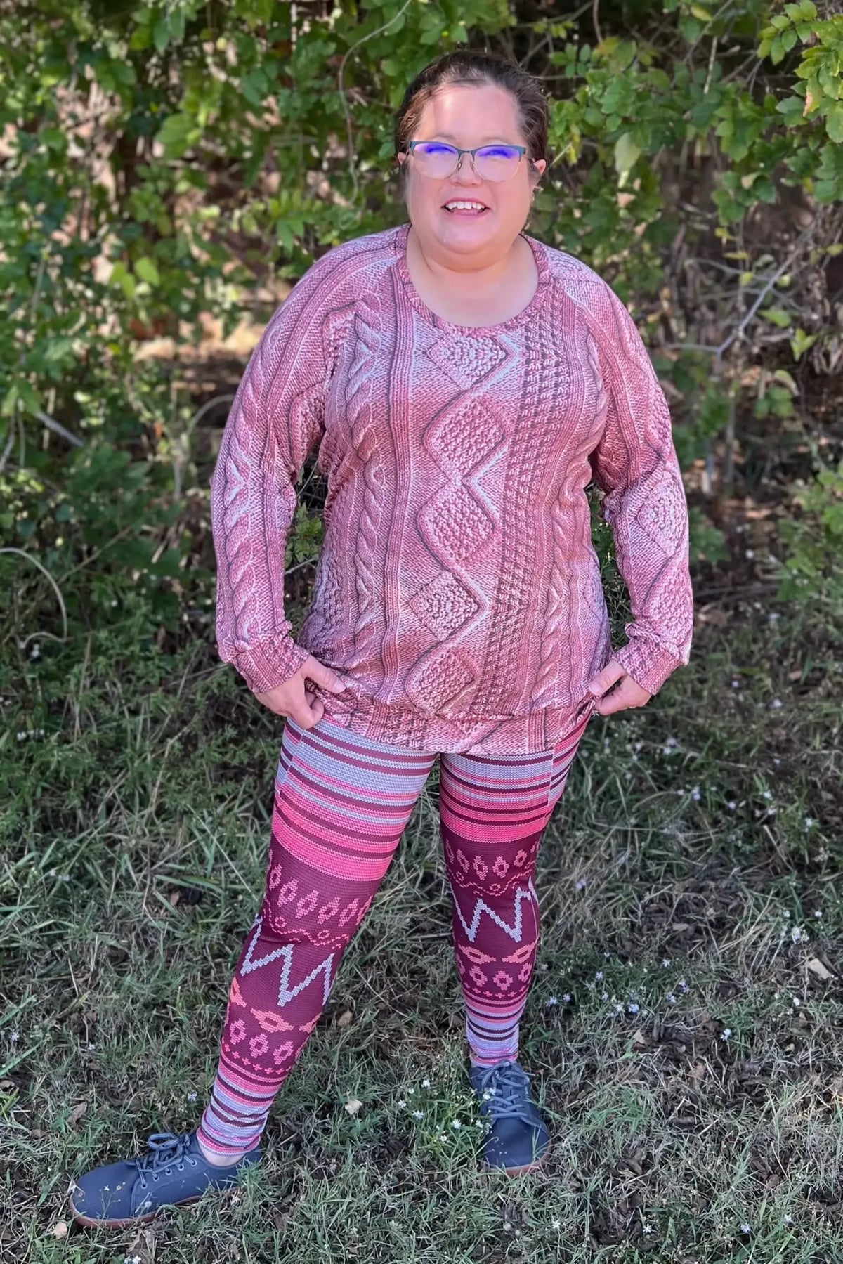 A woman in a rose cable knit top and patterned leggings stands outdoors.